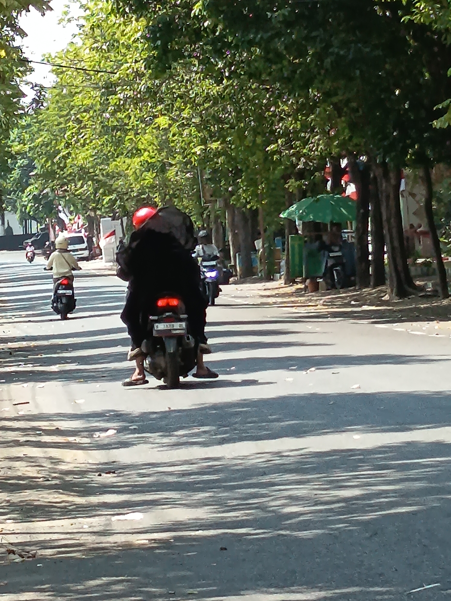 In Kandangjati Kulon, Indonesia, on a sunny day, a motorcyclist wearing a red helmet and a black hijab rides a dark-colored motorcycle. The rider is also wearing brown sandals. The motorcycle has a red taillight and a license plate. Another motorcyclist in a cream-colored jacket is visible further ahead on the left, also riding a dark motorcycle with a visible license plate. To the right, there is a green umbrella covering a street vendor's stall, with a person visible behind it, and another motorcycle parked nearby. The road is asphalt, and trees line the side of the road, casting shadows. Small pieces of debris are scattered on the road. In the background, there are buildings and more trees.