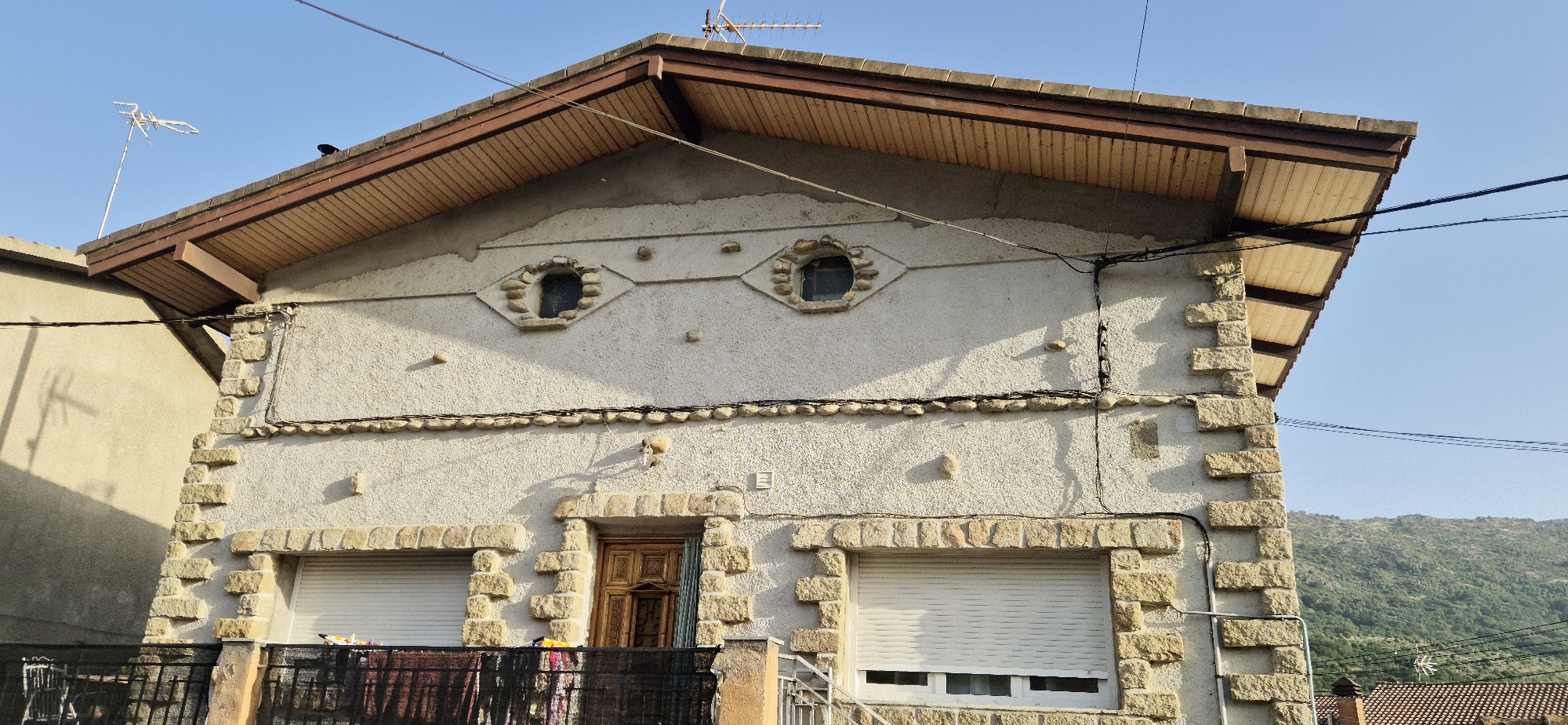 A daylight, eye-level, exterior shot shows the facade of a two-story, light-colored stucco house with a wooden-shingled overhang and a brown tiled roof. The house has a central wooden door flanked by two white shuttered windows. Above the windows are two small, circular, stone-framed windows arranged in a diamond pattern. The house is trimmed with large, rough-hewn, light brown stones on the corners and around the windows and door. Electrical wires run along the facade and across the roof. In the background, a green, tree-covered mountain range is visible under a clear blue sky. A television antenna is mounted on the roof to the left, and another antenna is visible on the mountain in the distance.