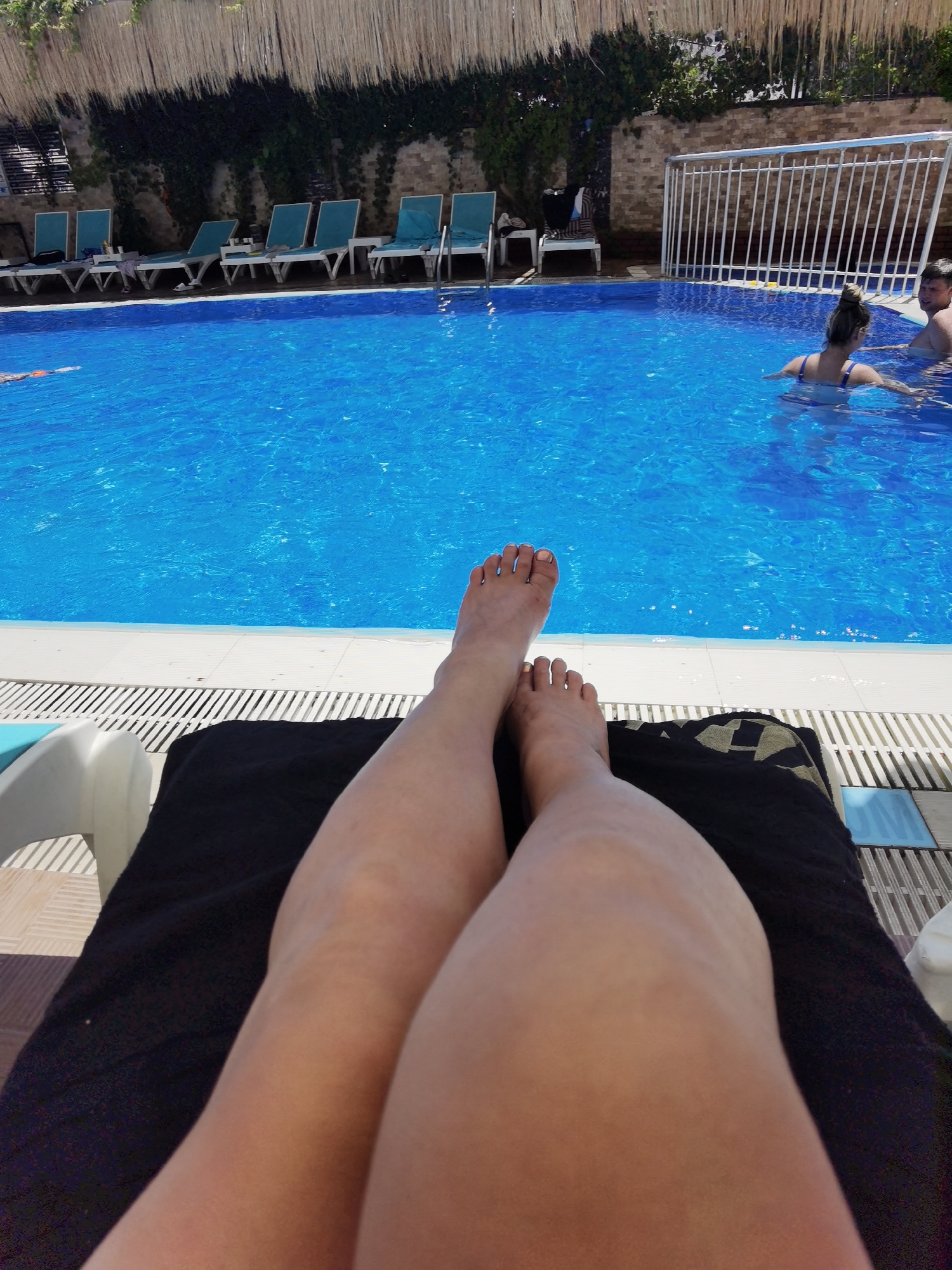 In Alanya, Turkey, a first-person perspective captures a scene at a poolside resort. The foreground is dominated by the legs and feet of an individual reclining on a dark-colored towel, placed on a sun lounger. The legs are bare and tanned, with painted toenails. The background reveals a vibrant blue swimming pool, with the water reflecting the sunlight. Several empty turquoise lounge chairs are neatly arranged along the edge of the pool. Further in the distance, two individuals are visible in the pool, one with a blonde bun and the other partially submerged. A white metal fence with a lattice design runs along the far side of the pool, adjacent to a brick wall covered in green ivy. Above the fence, a thatched-style roof structure is visible, suggesting an outdoor setting. The environment appears to be a sunny day, typical for a resort in Alanya.