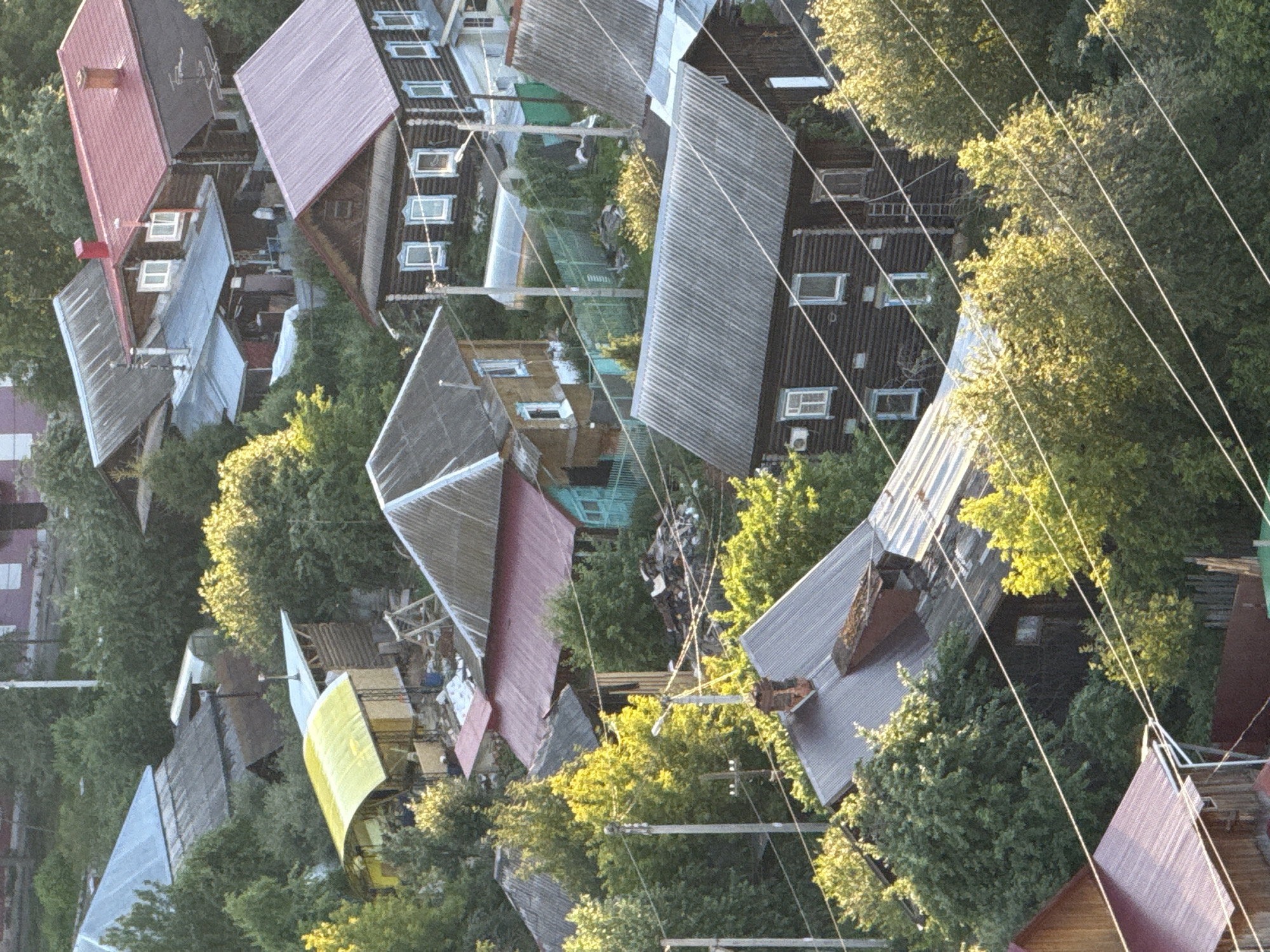 This aerial, eye-level, medium shot captures a collection of rural residential structures and dense foliage under natural daylight. The scene appears to be a village setting, possibly in Zarechnaya (Gamovskoe S/P), Russia, characterized by a mix of wooden and metal-roofed houses, some appearing older and weathered, others more recently constructed with colorful tin roofing. The architecture is predominantly traditional, featuring wooden log houses with prominent window frames and visible roofing materials like corrugated metal and asphalt shingles. Various outbuildings and sheds are interspersed within the property, some with arched, transparent roofs and wooden frames.
The environment is rich with mature trees and green vegetation, creating a sense of being surrounded by nature. Overgrown areas and piles of debris, including wood scraps and construction materials, suggest ongoing or recent activity. Power lines crisscross the scene, supported by wooden utility poles, indicating a functional residential area. No people are visible in the frame, and no specific actions or interactions are depicted. The overall impression is one of a lived-in, rural community with a blend of rustic charm and practical, everyday life. The lighting suggests it may be late afternoon or early morning due to the warm, directional light casting shadows.