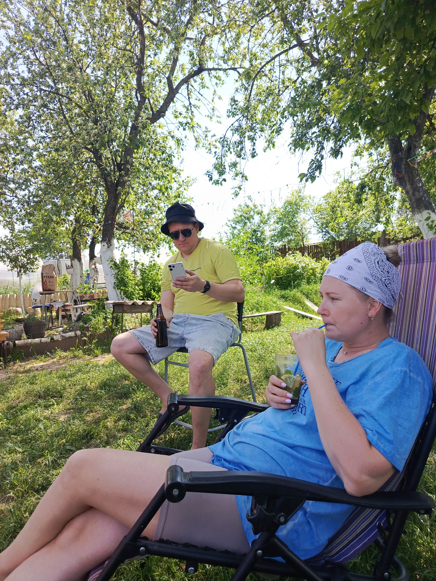 The image shows a man and a woman relaxing outdoors. The woman, wearing a blue t-shirt and a white patterned bandana, is seated in a reclining chair and drinking from a glass with a straw. The man, wearing a yellow t-shirt, blue shorts, sunglasses, and a black hat, sits on a chair, holding a bottle and looking at his phone. The scene takes place on a grassy lawn, with trees and other vegetation in the background. In the left background there is what appears to be a greenhouse and other garden-related objects. The location is likely the city of Chaikovsky, Russia.