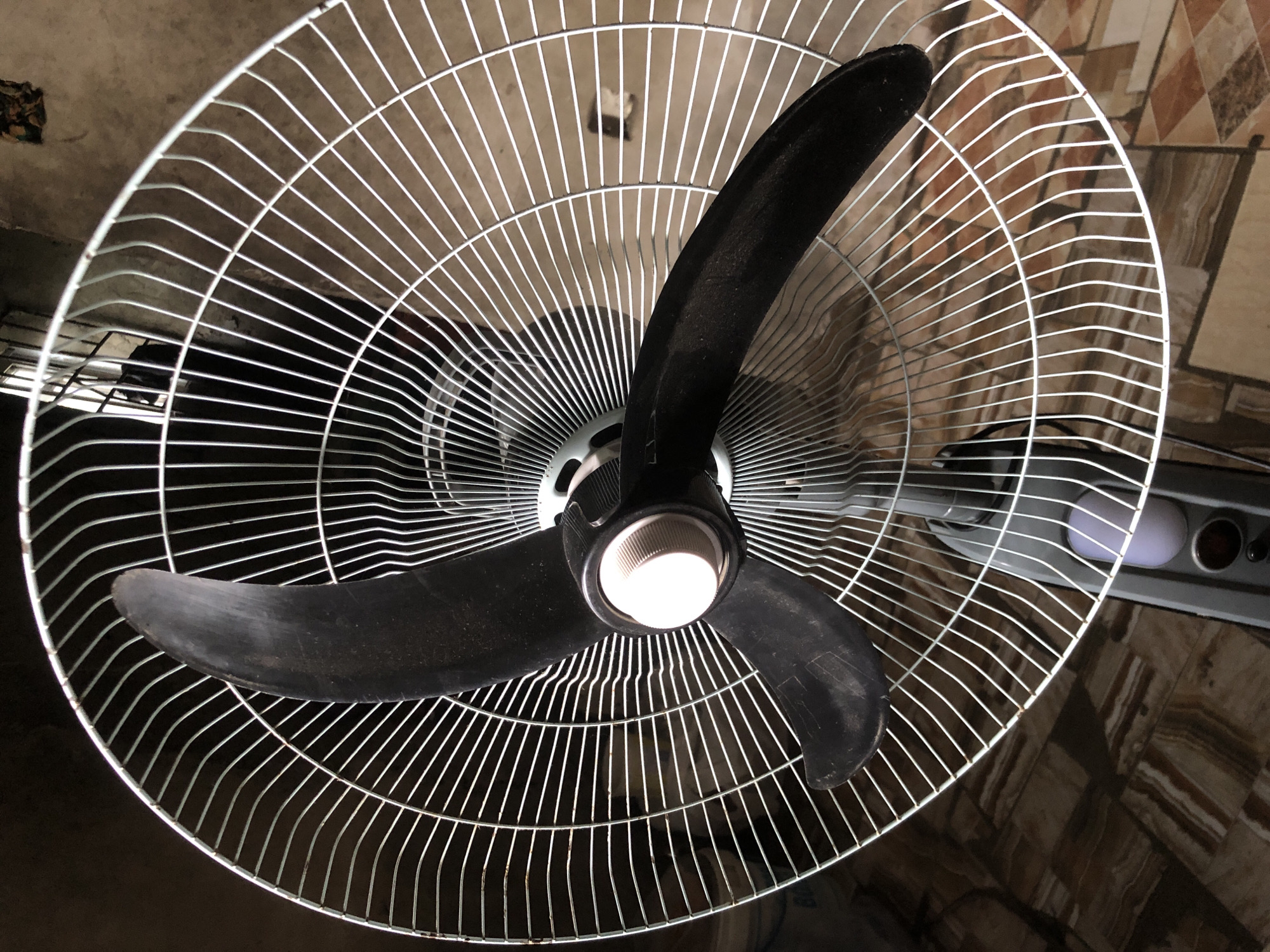 The image captures an upward view of a three-bladed ceiling fan in an indoor setting in Ado, Nigeria. The fan features a prominent white metal safety cage that is circular, encompassing the three dark, possibly black, fan blades. The blades appear stationary and show visible dust and wear. The central hub of the fan, from which the blades extend, is black with a lighter, possibly white or off-white, conical cap in its center.
The fan is mounted on a light-colored, seemingly concrete ceiling, which exhibits some imperfections and minor damage in certain areas. Through the fan's cage, particularly in the background to the right and bottom right, patterned floor tiles in shades of brown, beige, and terracotta are visible, suggesting an existing floor or perhaps an adjacent tiled wall. To the right side of the fan, partially obscured by the fan's cage, is a section of a grey appliance or device, possibly an air conditioning unit or another fan's motor, featuring what appears to be a light-colored, possibly purple or white, indicator light and another darker, reddish-brown circular element. The overall lighting suggests it is daytime or a well-lit indoor space. No people or visible text are present in the image.