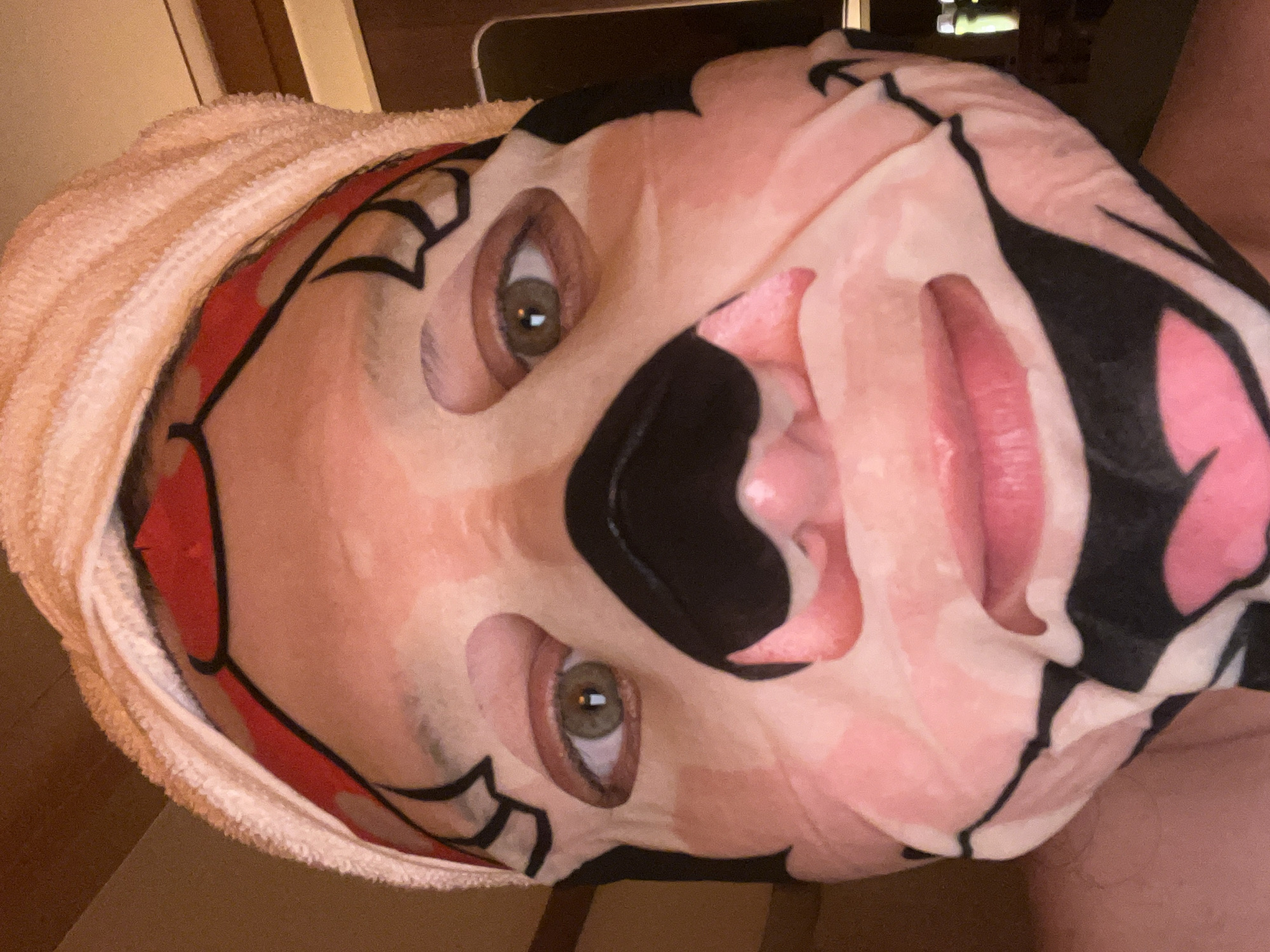 A person wearing a sheet mask with a cartoon design, consisting of pink, black, and white colors. The mask covers the majority of their face, but cutouts expose their eyes and mouth. A towel is wrapped around their head. There is no indication of location, including the city of Tel-Aviv, Israel.