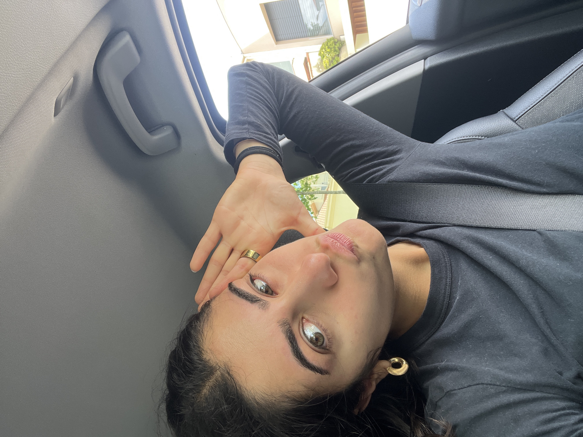 A woman is taking a selfie inside a car. She is wearing a black long-sleeved shirt and a gold ring and earring. She is wearing a seatbelt and holding her head with her hand. Outside the car window, a building with plants can be seen. Based on the architecture, the location is likely Porto, Portugal.