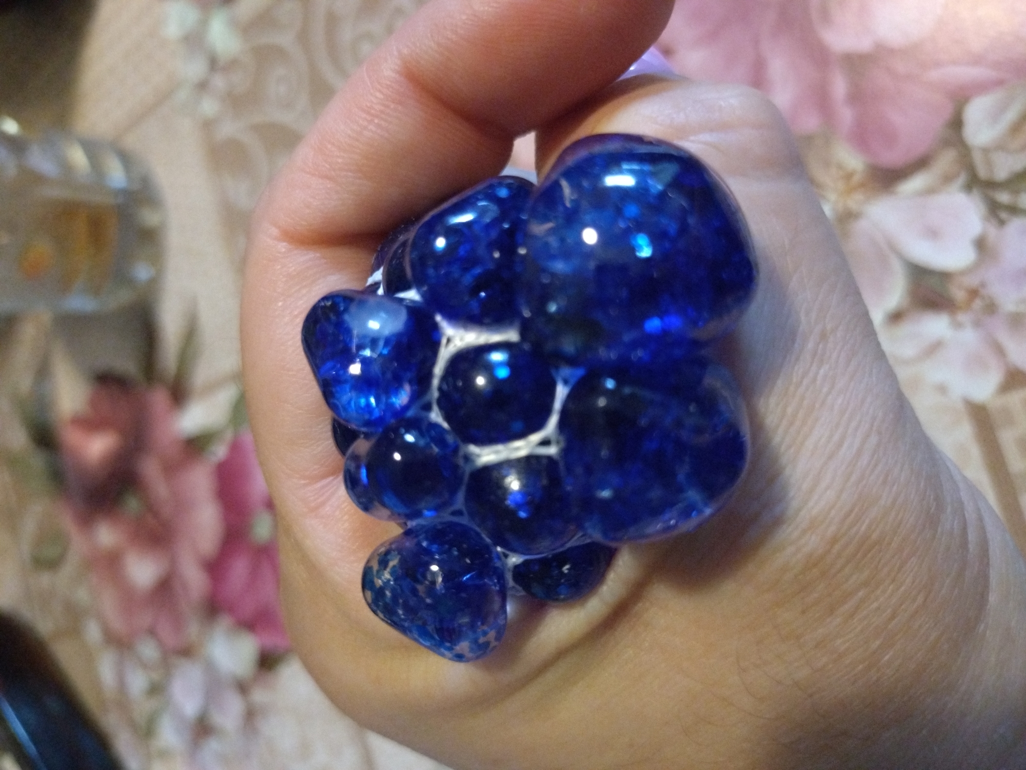 A hand is holding a blue squishy ball filled with smaller, iridescent blue balls. The ball is made of a netting material that contains numerous small, smooth, round, blue spheres, each reflecting light. The background is a blurred surface with a floral pattern in shades of pink and white, possibly a tablecloth. The lighting suggests an indoor setting. There is no information in the image to indicate its location in Stara Zagora, Bulgaria.