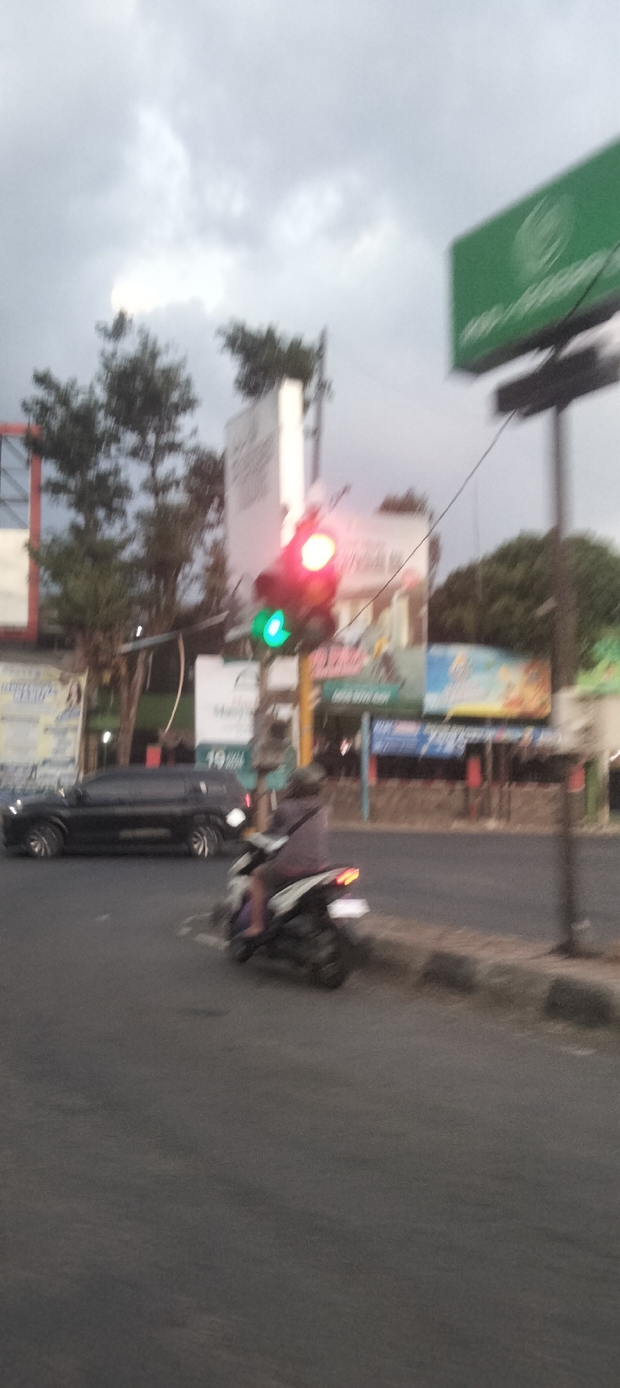 In Patrang, Indonesia, a street scene unfolds under a cloudy sky. A traffic light, with the red and yellow lights illuminated, stands at an intersection. A black SUV is captured mid-motion on the left side of the frame, while a person on a scooter, wearing a helmet and a dark top, is moving away from the camera in the right foreground. Billboards and buildings are visible in the background, suggesting an urban environment. The image appears to have been taken during daylight hours, likely late afternoon or early evening due to the diffused light and cloudy conditions.
