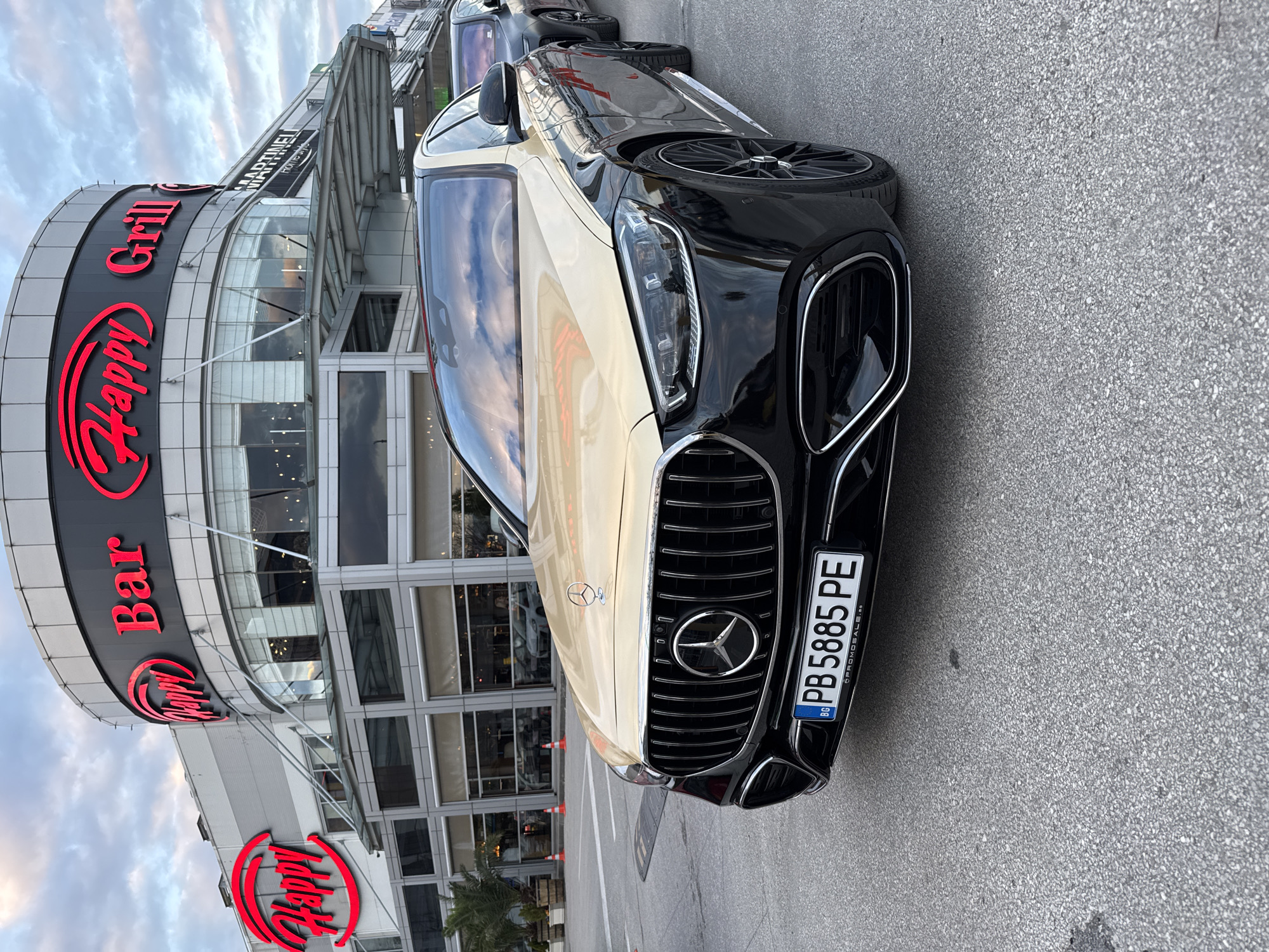 The image displays a two-tone Mercedes-Benz sedan parked on an asphalt surface in front of the Happy Bar & Grill restaurant in Plovdiv, Bulgaria. The building, identified by prominent red neon signage spelling "Happy Bar & Grill" and partially "MARTINELI HOME STYLE," features a modern, multi-story design with a curved glass facade. The primary vehicle, a Mercedes-Benz, exhibits a black finish on its lower body and a gold/champagne color on its hood, roof, and upper body panels. Its front grille features vertical slats and a central Mercedes-Benz emblem. The visible license plate reads "PB 5885 PE". A second, dark-colored sedan is partially visible parked directly behind the Mercedes-Benz. The immediate environment includes grey asphalt pavement, a speed bump, several small palm trees in planters to the left, and distant orange traffic cones. The sky is overcast with scattered clouds, indicating daylight conditions. No individuals are discernible in the scene.
