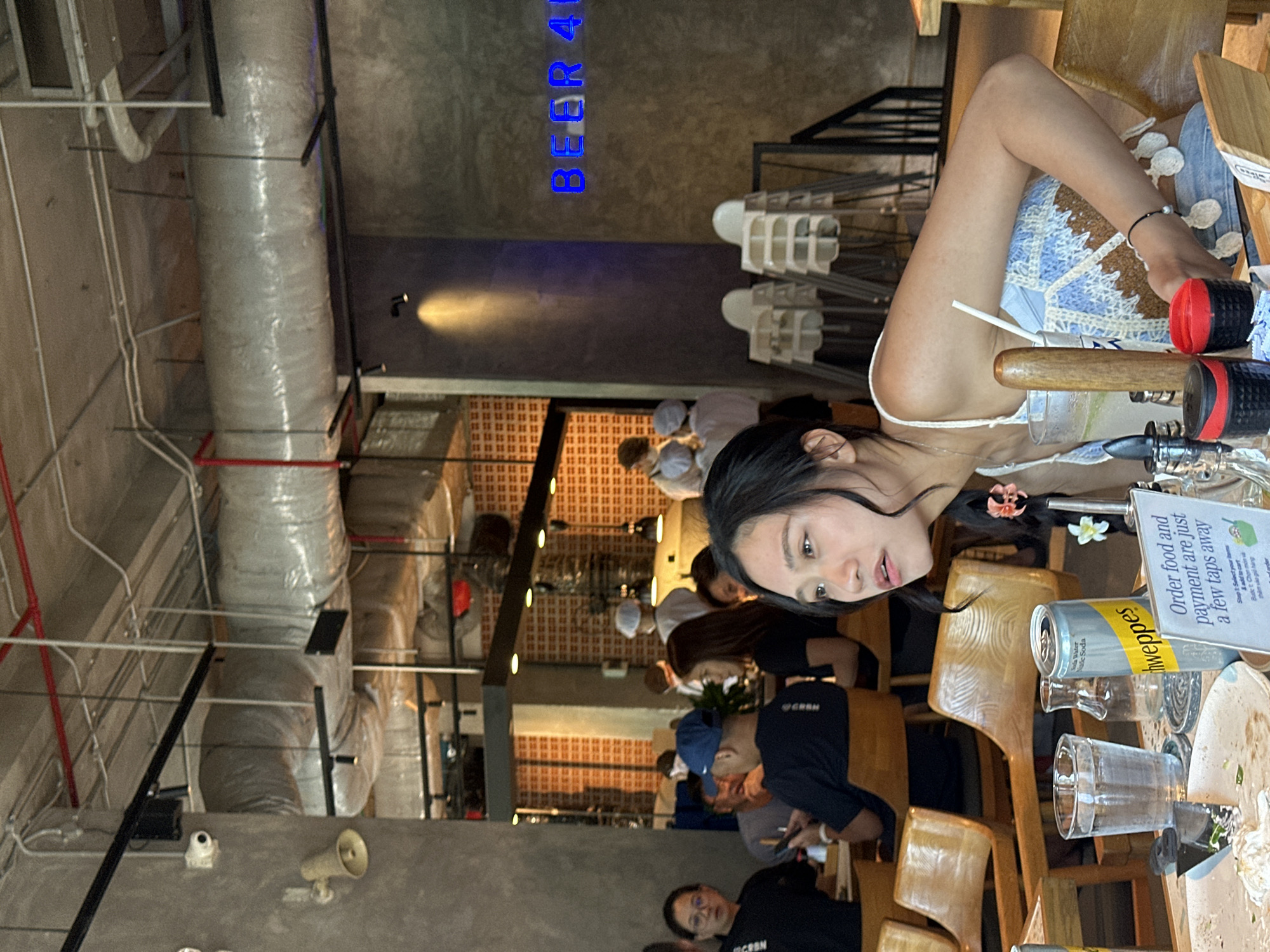 This image shows a group of people inside a restaurant with an industrial-chic design, featuring exposed concrete walls, pipes, and ductwork. The lighting is dim, with spotlights highlighting various areas. In the foreground, a young woman with dark hair, wearing a blue and white crochet top, is looking to her right. In front of her is a table with drinks, including a glass of water and a can of Schweppes soda, as well as some food remnants on a plate. Other patrons are visible in the background, some of whom appear to be staff members wearing chef hats. A neon sign in blue lights on the wall reads "BEER 4U". The restaurant appears to be located in Da Nang, Vietnam, a city known for its vibrant culinary scene and modern establishments.