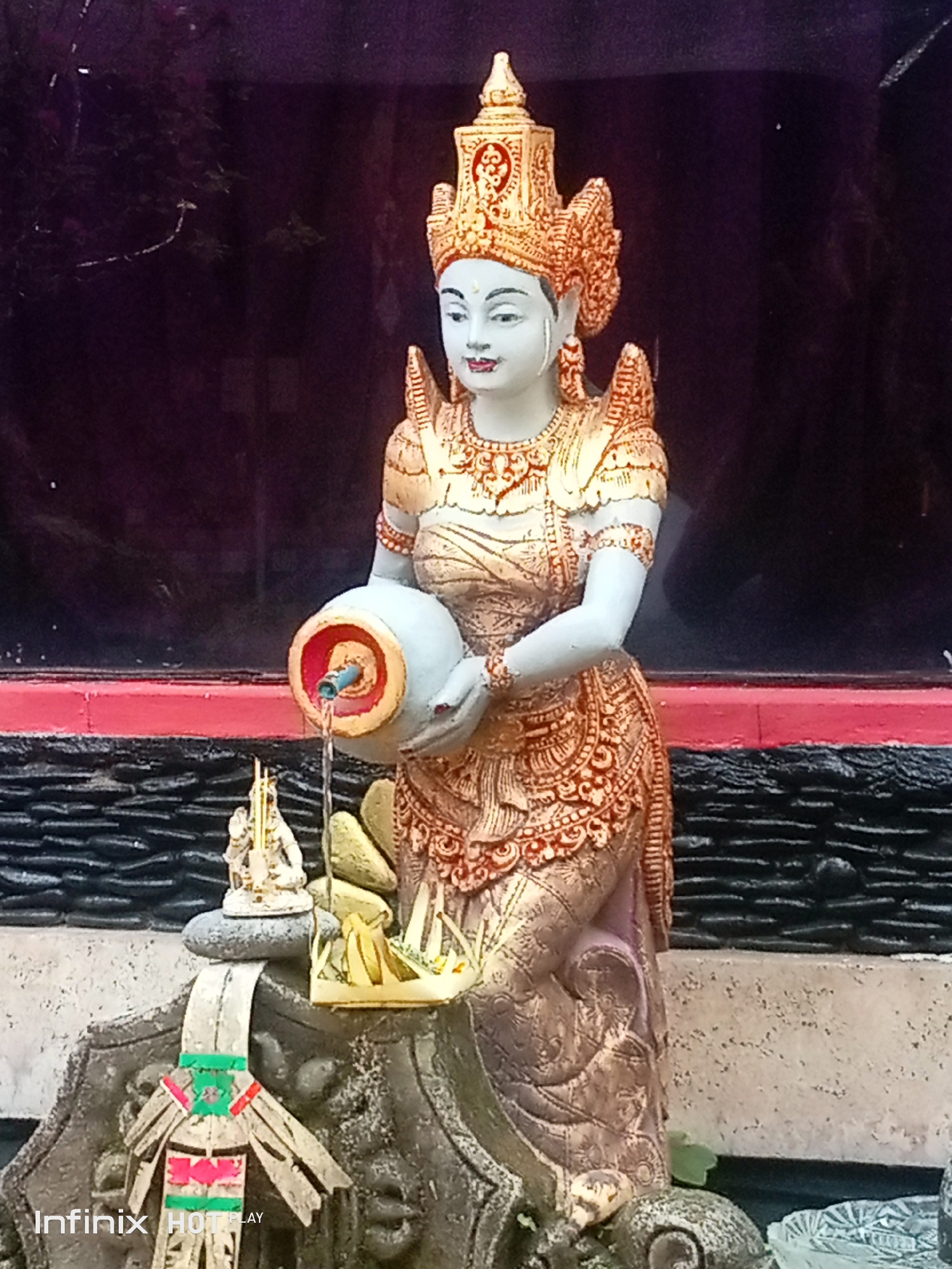 A statue of a woman in ornate golden attire, wearing a crown, stands in what appears to be a shrine or outdoor temple setting. The statue is holding a light-colored jug from which water is flowing, creating a small fountain effect. Below the statue, on a stone base, are several smaller offerings or decorative items, including a tiny statue of a figure with arms raised, and some golden objects. The background consists of dark, reflective surfaces and a red band. The statue is positioned in front of a textured, dark gray wall. The scene is likely located in Blahbatuh, Indonesia, a region known for its artistic traditions.