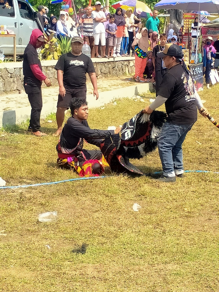 The image depicts an outdoor cultural performance or preparation for one, occurring on a grassy field in Celep, Indonesia. In the center, a male individual wearing a black long-sleeved shirt and a colorful patterned sarong with red, yellow, and purple elements is seated on the grass, facing right. He is interacting with a large, elaborate, animal-like costume or mask, predominantly black with white and red patterns and black and white feathery accents. Another male, wearing a black t-shirt, blue jeans, and a cap, stands to the right of the seated individual, holding the rear section of the costume and a wooden pole attached to it, appearing to assist.
To the left of the central interaction, two other males stand observing. One, in a black t-shirt and dark shorts, stands slightly behind the seated performer. The other, wearing a red-hooded long-sleeved shirt under a black t-shirt and dark pants, stands further left.
In the background, a crowd of spectators, including adults and children, watches from an elevated concrete platform behind a low stone wall. Many individuals are wearing casual clothing, and several women wear headscarves. To the far left, a light-colored van is parked. To the right background, several market stalls or tents with colorful umbrellas are visible, one displaying "Bintang SF" on its canopy. The ground is covered in dry, light green grass with patches of dirt. A light blue cord or rope stretches across the grass in the lower half of the frame, and a few discarded plastic bottles are scattered on the ground. The scene is brightly lit by natural sunlight, indicating clear weather.