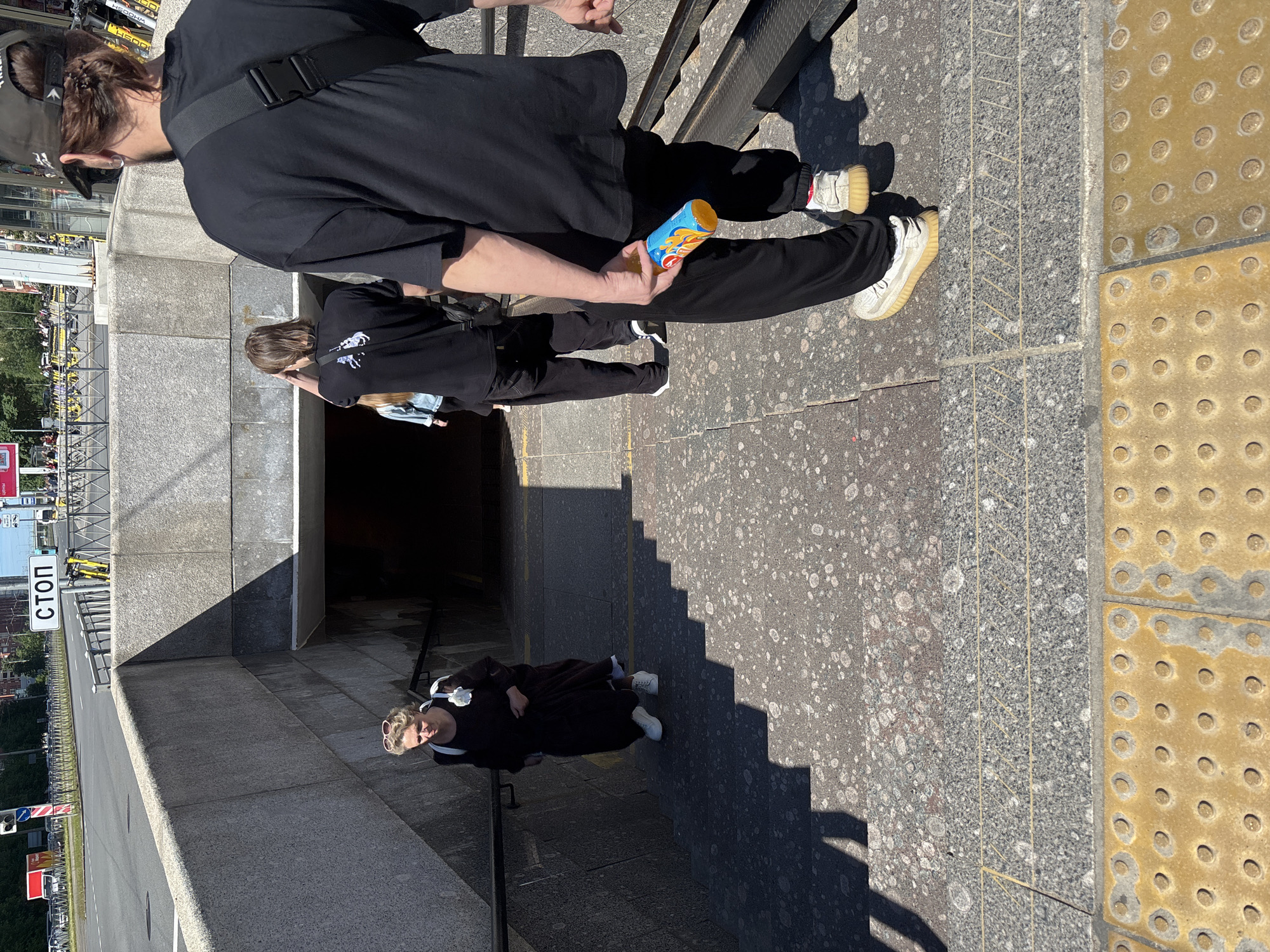 In an outdoor urban setting, three individuals are descending a set of granite stairs leading into a subway entrance. The lighting suggests a sunny day, casting distinct shadows. The stairs are wide and appear to be made of speckled granite, with a tactile paving system of yellow circles at the bottom.
On the left, a woman with blonde hair and sunglasses on her head walks down the stairs. She is wearing a long, dark dress and white sneakers, carrying a white bag over her shoulder.
Further down the stairs, two individuals are walking in the same direction. The person in front has medium-length brown hair and is wearing a black t-shirt with a white graphic on the back, black pants, and dark sneakers. They are carrying a black bag across their body. The person behind them, facing away from the camera, is wearing a black baseball cap, a black t-shirt, and black pants. They have a black bag slung over their shoulder and are holding a blue and orange can in their right hand. Their sneakers are light-colored with a thick sole.
The subway entrance is visible in the background, appearing dark and recessed. Above the entrance, a sign with the Cyrillic word "СТОП" (STOP) is mounted on a metal railing. In the distance, beyond the stairs and railing, a street with traffic and a red truck is visible, along with some parked yellow bicycles. The overall environment appears to be a public thoroughfare in a city.