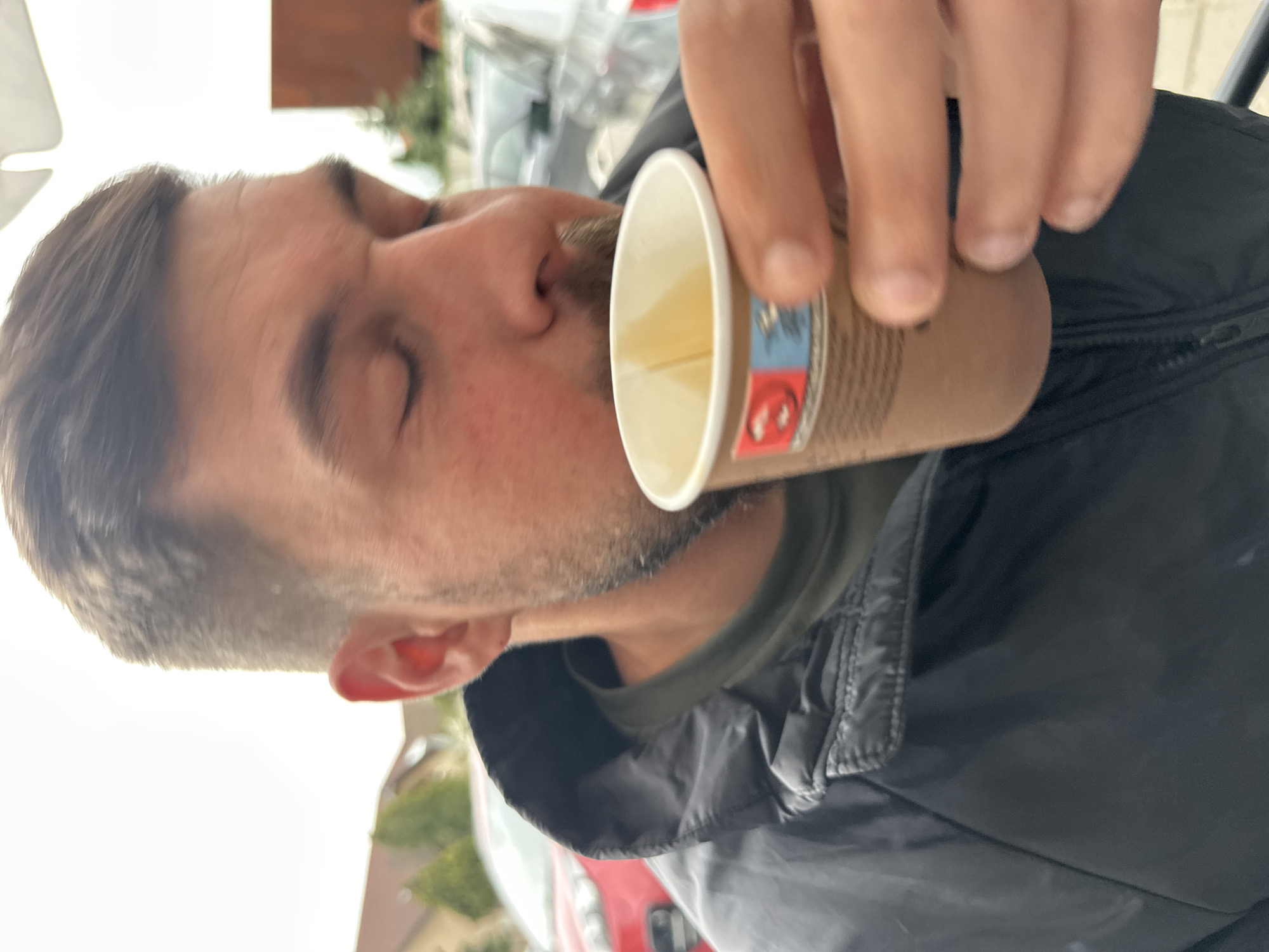 The image shows a close-up, profile view of a man drinking from a disposable paper cup in an outdoor setting in Plovdiv, Bulgaria. The man, seen from the mid-chest up, faces left with his eyes closed. He has short brown hair and light stubble or a short beard, and is wearing a dark green t-shirt under a black zip-up jacket.
His right hand holds a brown paper cup with a white rim, containing a light yellow liquid, possibly tea or juice. The cup features a small, colored design on its side with blue and red sections, though no clear text or detailed logos are discernible.
The background is blurred, suggesting an outdoor environment during daytime. Vague shapes of vehicles, including a red car, and some greenery are visible in the soft-focus background, alongside a bright, possibly overcast sky or light-colored building facade. The man's closed eyes might convey concentration or enjoyment while he takes a sip from the cup.
