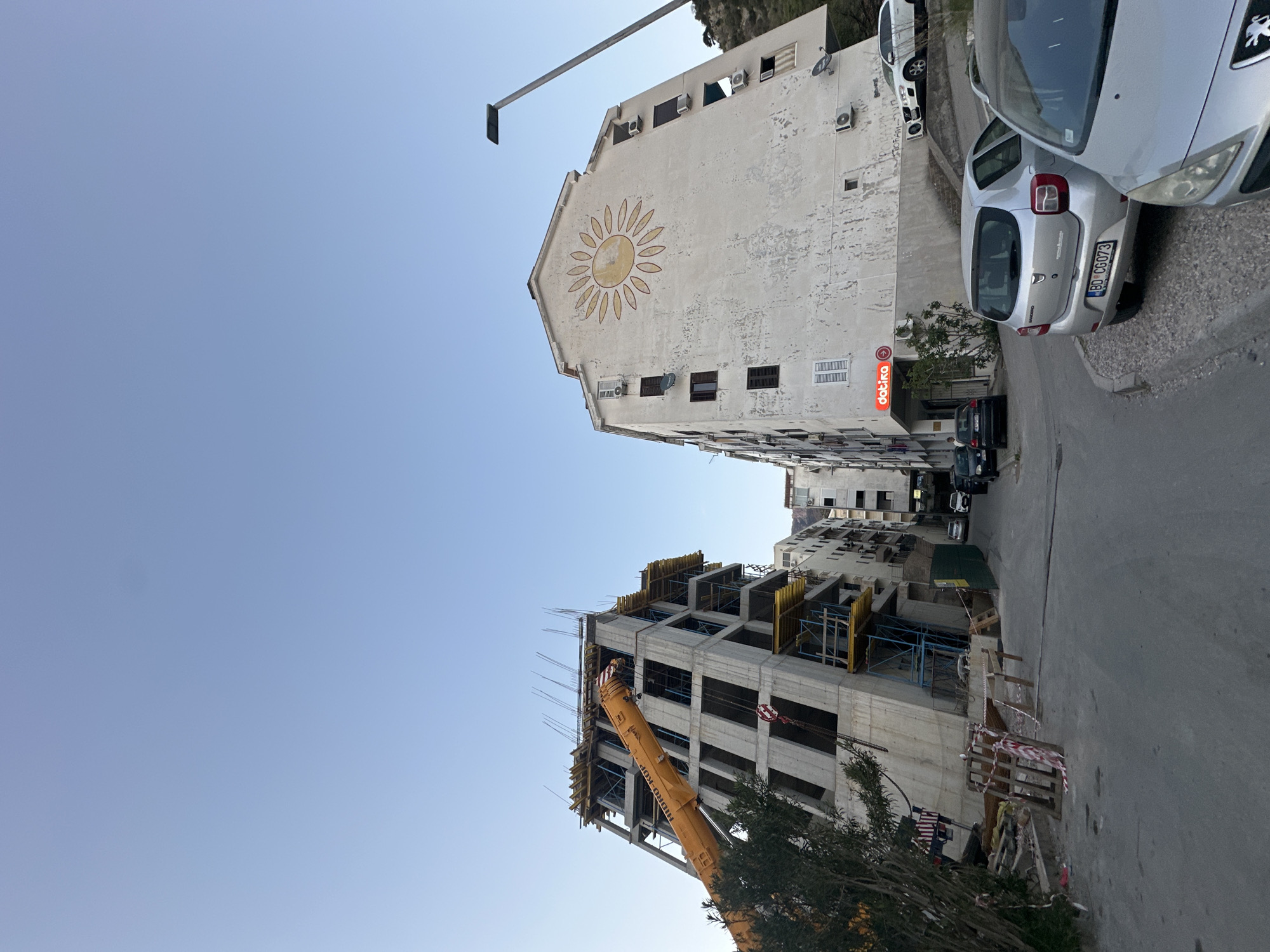 An eye-level shot depicts a street in Budva, Montenegro, bordered by buildings and a construction site. The sky is clear and blue. The buildings vary in architectural style and condition. One older building features a faded mural of a sun. A new building is under construction, with scaffolding and a crane prominently visible. Several cars are parked along the street. The street itself appears paved. A datika sign is visible on one of the buildings.