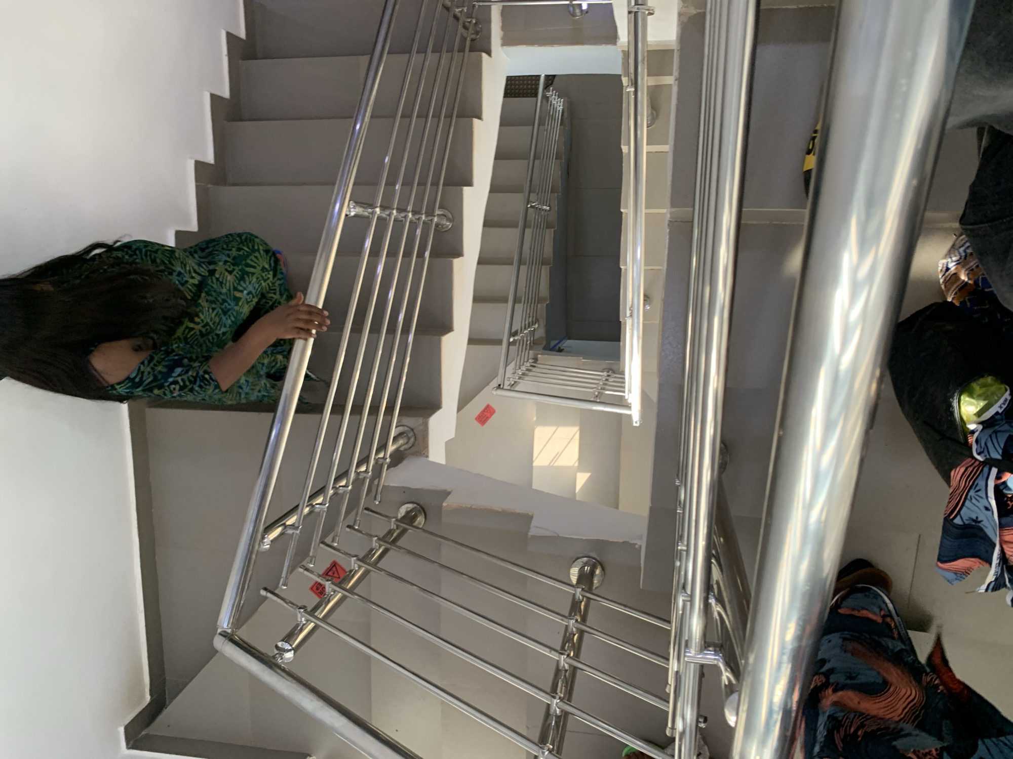 The image depicts a person descending a well-lit, multi-story staircase. The staircase features modern, polished metal railings and grey tiled steps. The person, viewed from above, is wearing a green and blue patterned off-the-shoulder dress and has long, dark hair. Sunlight casts shadows on the stairs, indicating an outdoor or well-lit interior space. A red hazard sticker is visible on one of the stair risers. The overall environment suggests a contemporary building, possibly a residential complex or commercial space. The location is identified as Jos, Nigeria.