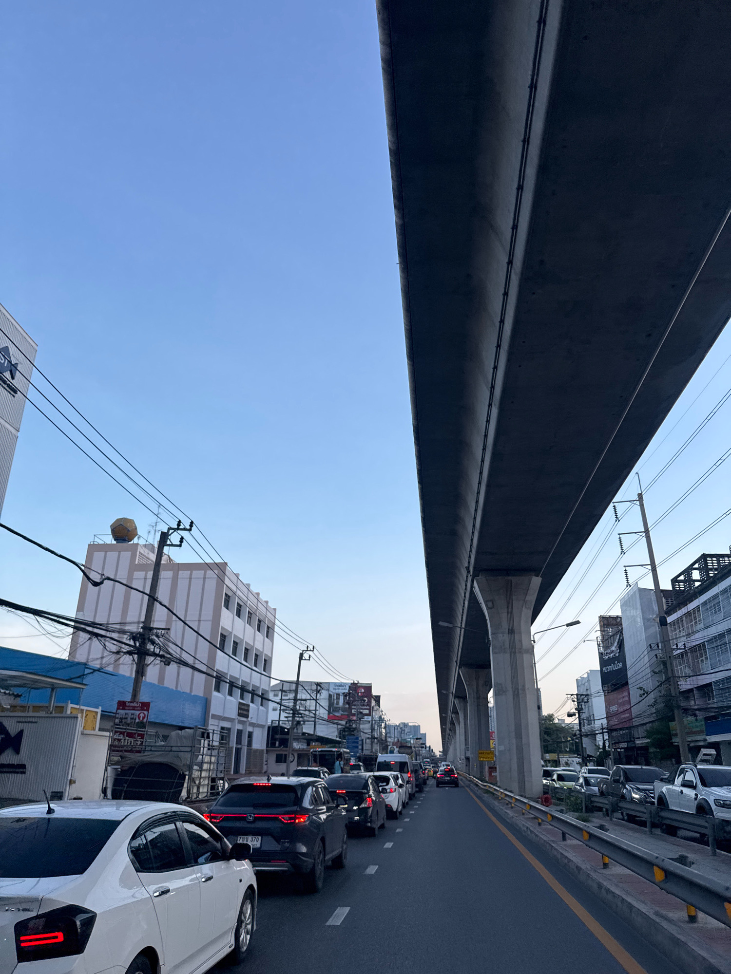 The image depicts a street scene in Samrong Nuea Subdistrict Municipality, Thailand, characterized by heavy traffic congestion under an elevated highway structure. The sky is clear and pale blue, suggesting either early morning or late afternoon. The elevated highway, constructed of concrete and supported by large pillars, dominates the upper portion of the frame.
The street below is filled with numerous vehicles, primarily cars and some trucks, forming a long queue in both visible lanes. The vehicles appear to be stationary or moving very slowly. Among the visible cars are a white sedan in the foreground, a black SUV, and other vehicles further down the road. Many of these vehicles have their brake lights illuminated, indicating the traffic jam.
On either side of the street are multi-story buildings, typical of an urban environment. These buildings display various signs and advertisements, some in Thai script. Electrical wires and utility poles are prominently visible, crisscrossing above the street and along the buildings. One building on the left features a large, spherical, golden object on its rooftop, possibly a weather balloon or decorative element.
The environment appears to be a commercial or mixed-use area within the city, with the elevated highway indicating a major transportation artery. The overall impression is one of a busy, developed urban area experiencing significant traffic.