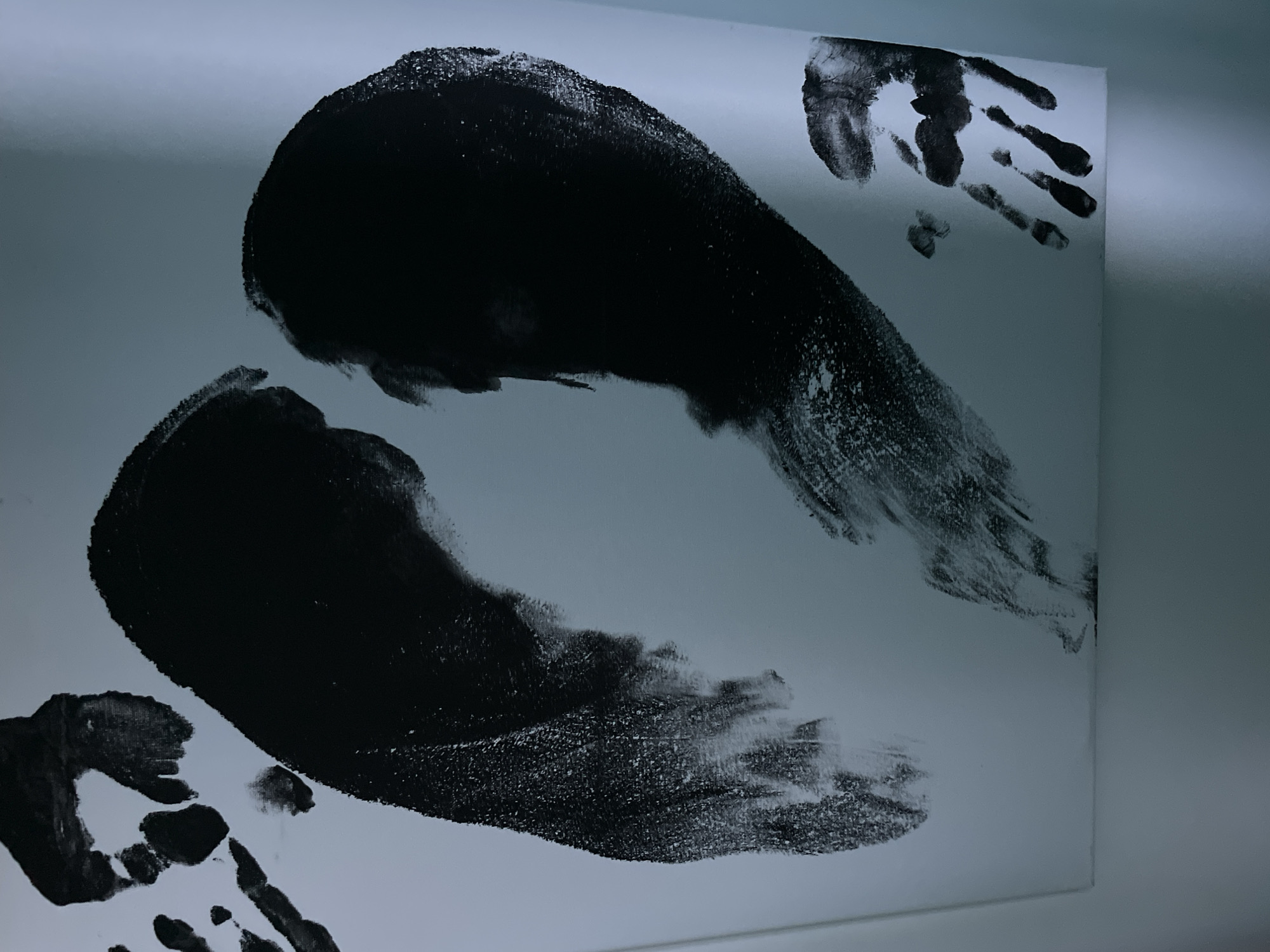 A close-up shot depicts a white canvas board mounted on a light gray wall. The canvas features dark, black paint handprints. Two larger, amorphous blots dominate the central area, with smaller, distinct handprints appearing at the top right and bottom left corners. The painting is located in an unknown city, with an unknown address.