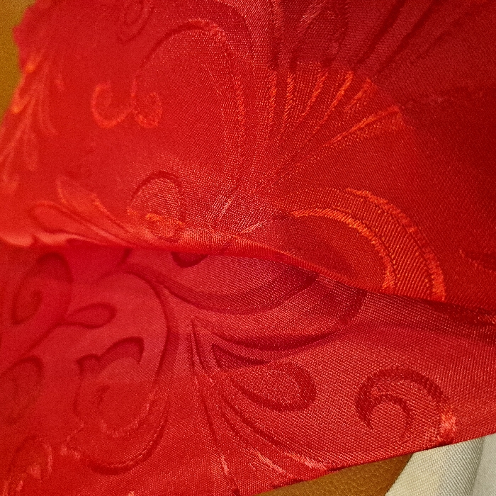 The image showcases red fabric with an intricate, embossed floral pattern. The fabric has a soft, shiny texture, possibly silk or satin. A fold in the material creates depth and shadows, enhancing the visibility of the design. A glimpse of golden yellow and white fabrics appears at the bottom edge, suggesting a layered composition.