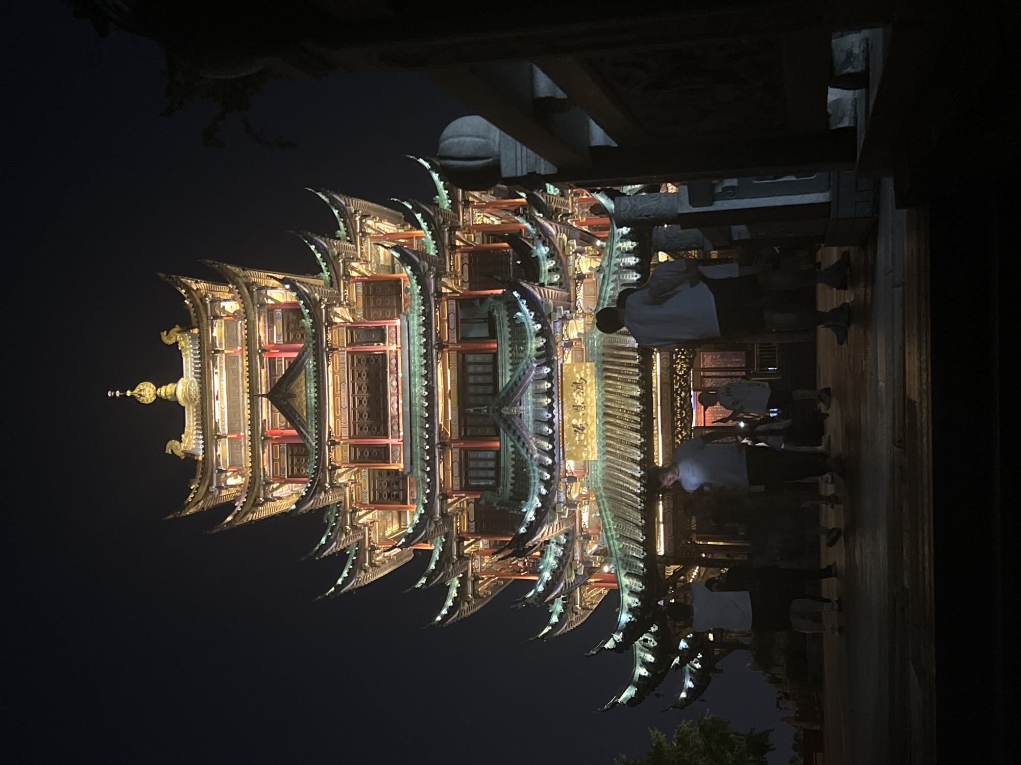 The image captures the Yellow Crane Tower at night, a prominent landmark in Wuhan, Hubei province, China, situated on the eastern bank of the Yangtze River. The tower, illuminated with colorful lights, features multiple tiered roofs adorned with intricate carvings and traditional Chinese architectural elements. A group of people, appearing to be tourists, are visible in the foreground, some standing and others walking, observing the illuminated structure. The dark night sky provides a dramatic backdrop, highlighting the vibrant lights of the tower. The foreground shows a paved area, likely a viewing platform or pathway, with some structural elements of a balcony or railing to the right. The overall scene depicts a well-lit tourist attraction at night, with visitors present.