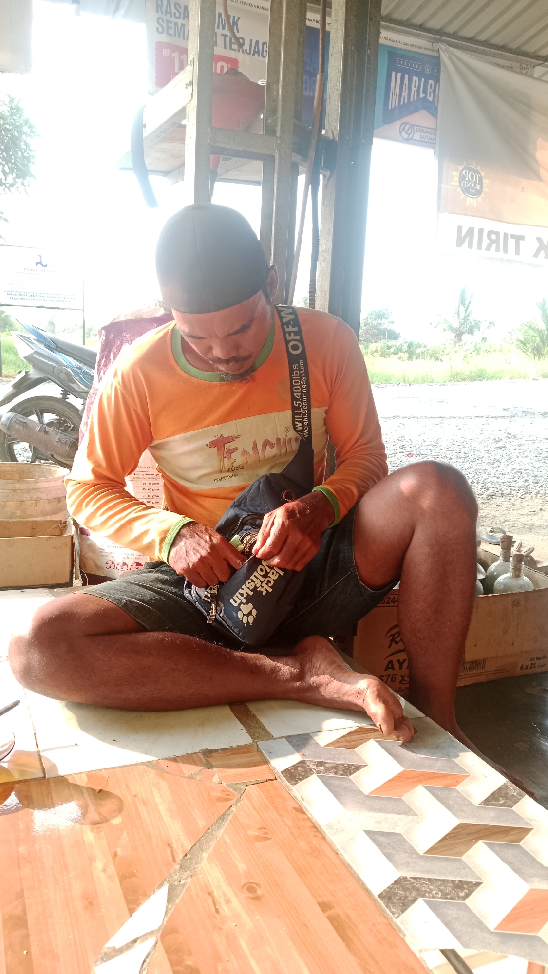 A man wearing an orange long-sleeved shirt and a dark cap is sitting cross-legged on the floor of an open-air structure. He is looking down and appears to be searching through a dark shoulder bag with the brand "Jack Wolfskin" visible. The bag is worn across his chest. He is wearing denim shorts. The floor is tiled with a pattern of interlocking geometric shapes in shades of brown, white, grey, and orange. To his left, there is a motorcycle and some sacks. To his right, a cardboard box is visible containing several glass bottles with green liquid. In the background, there are banners with text in Indonesian, including one that appears to be an advertisement for Marlboro. The scene is brightly lit, suggesting it is daytime, with sunlight streaming in from the left. The environment appears to be outdoors, possibly a roadside stall or a workshop in Karang Indah, Indonesia, given the banners and general setting.
