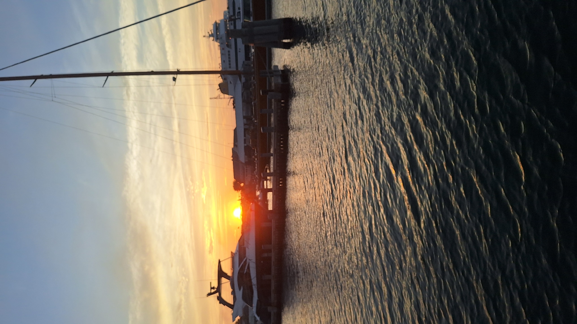 The image depicts a sunset over the water in Nantucket, United States of America. The sun, a bright orange orb, is low on the horizon, casting a warm glow across the sky and reflecting on the rippling water. Several boats are docked in the foreground, including a sleek white yacht and a larger white ferry or ship in the background. A tall mast from a sailboat is visible on the right side of the frame, with its rigging silhouetted against the colorful sky. Buildings are visible along the shore behind the boats, appearing as dark silhouettes against the sunset. The water is dark with streaks of light reflecting from the sun. The overall scene conveys a peaceful and serene atmosphere of a harbor at dusk.