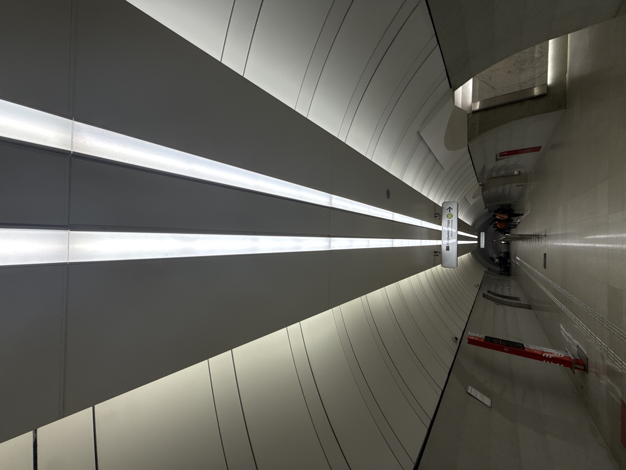 The image depicts the interior of a Moscow Metro station in Russia. The perspective is from the side of the platform, showing a long, curving tunnel-like space with a modern design. The walls are a light, neutral color, with horizontal lines running along their length, giving a sense of depth. There are two parallel light strips on the ceiling which are the light source. At the end of the tunnel, several people are seen. There are red signage on the right wall.