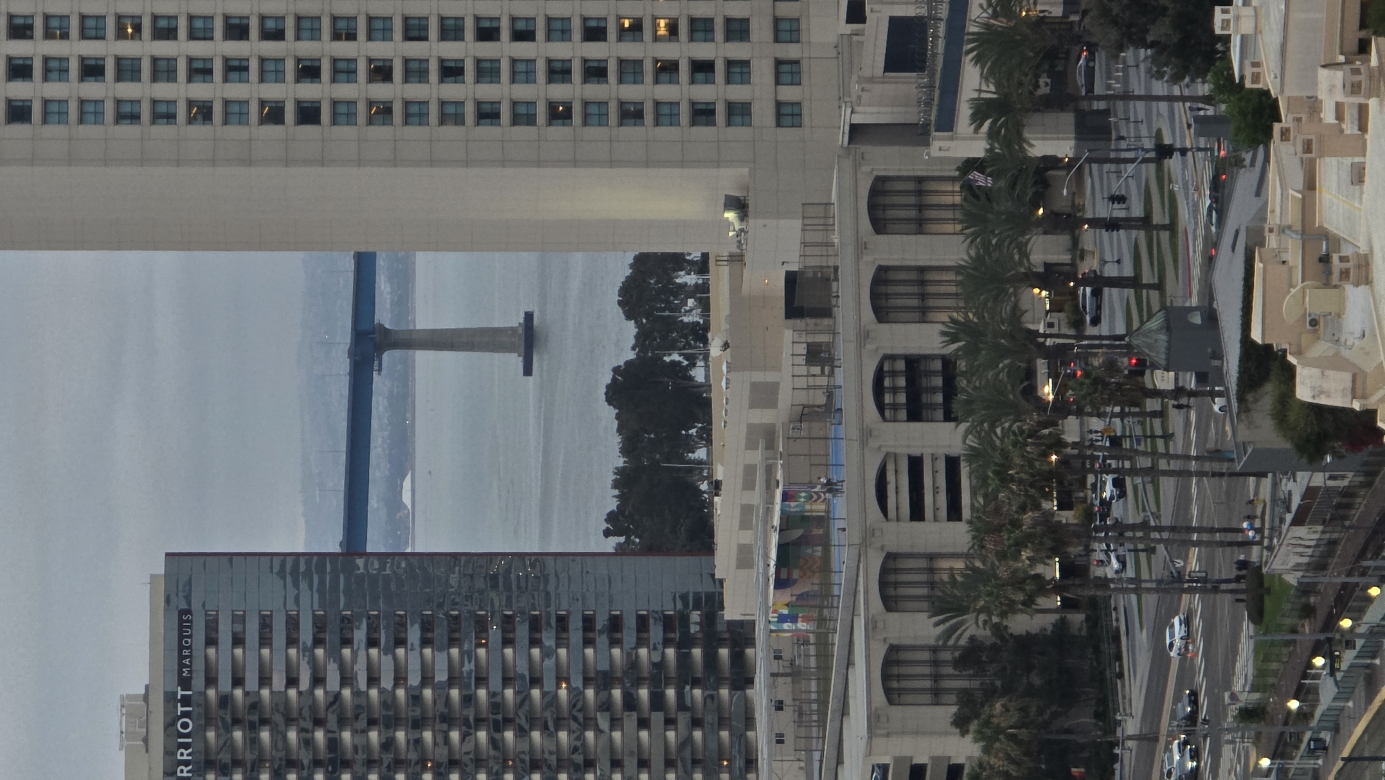 The image depicts an aerial view of the San Diego Bay, with the Coronado Bridge prominently featured in the background. To the left, the Marriott Marquis San Diego Marina hotel is visible, its modern architecture contrasting with the bridge. To the right, another large hotel building rises, its facade lined with windows. In the middle ground, a lower-rise building with a colorful mural on its roof is situated, adjacent to an outdoor recreational area. The foreground reveals a street scene with palm trees, traffic, and pedestrians, indicating an urban environment. The overall atmosphere is subdued, possibly due to overcast weather or the time of day.