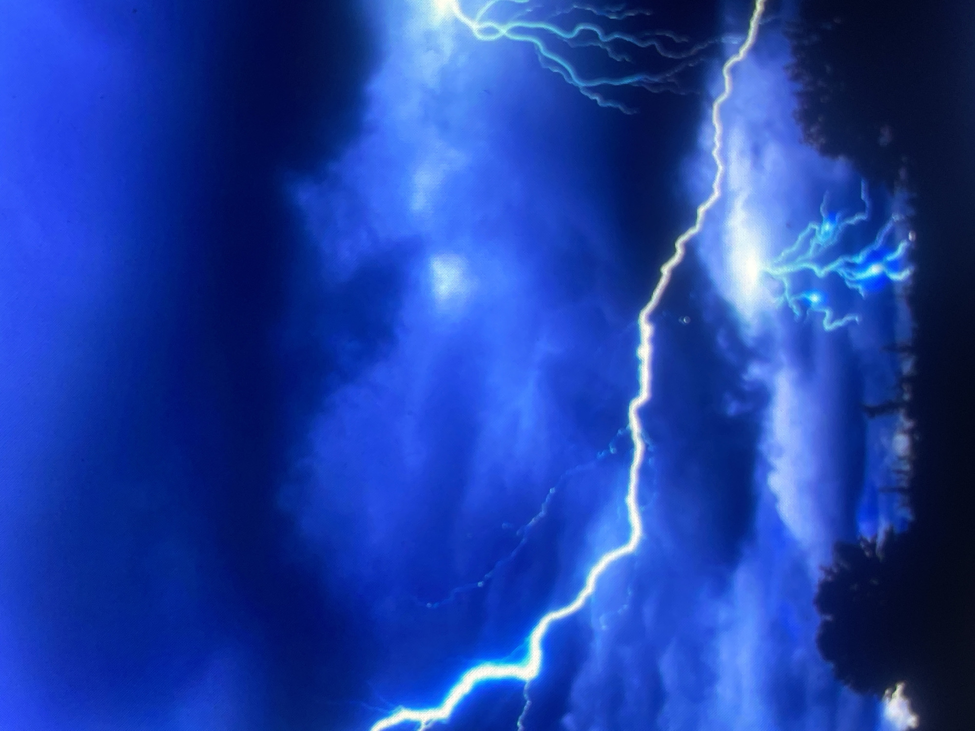 The image captures a stormy, nocturnal scene over Zelenyi Hai, Ukraine. The sky is dominated by deep blues and dark grays, indicative of heavy cloud cover during a thunderstorm. Multiple lightning strikes are visible across the frame. A prominent, bright white to electric blue lightning bolt extends from the upper left corner, diagonally across the middle of the image, curving towards the bottom-center. On the far right, a branched, electric blue lightning strike illuminates the clouds. A third, similarly branched electric blue bolt is present near the bottom-center, appearing to strike the ground. The lower third of the image features a dark, silhouetted horizon line, composed of dense foliage and trees against the illuminated sky. No human subjects or discernible man-made structures are present.