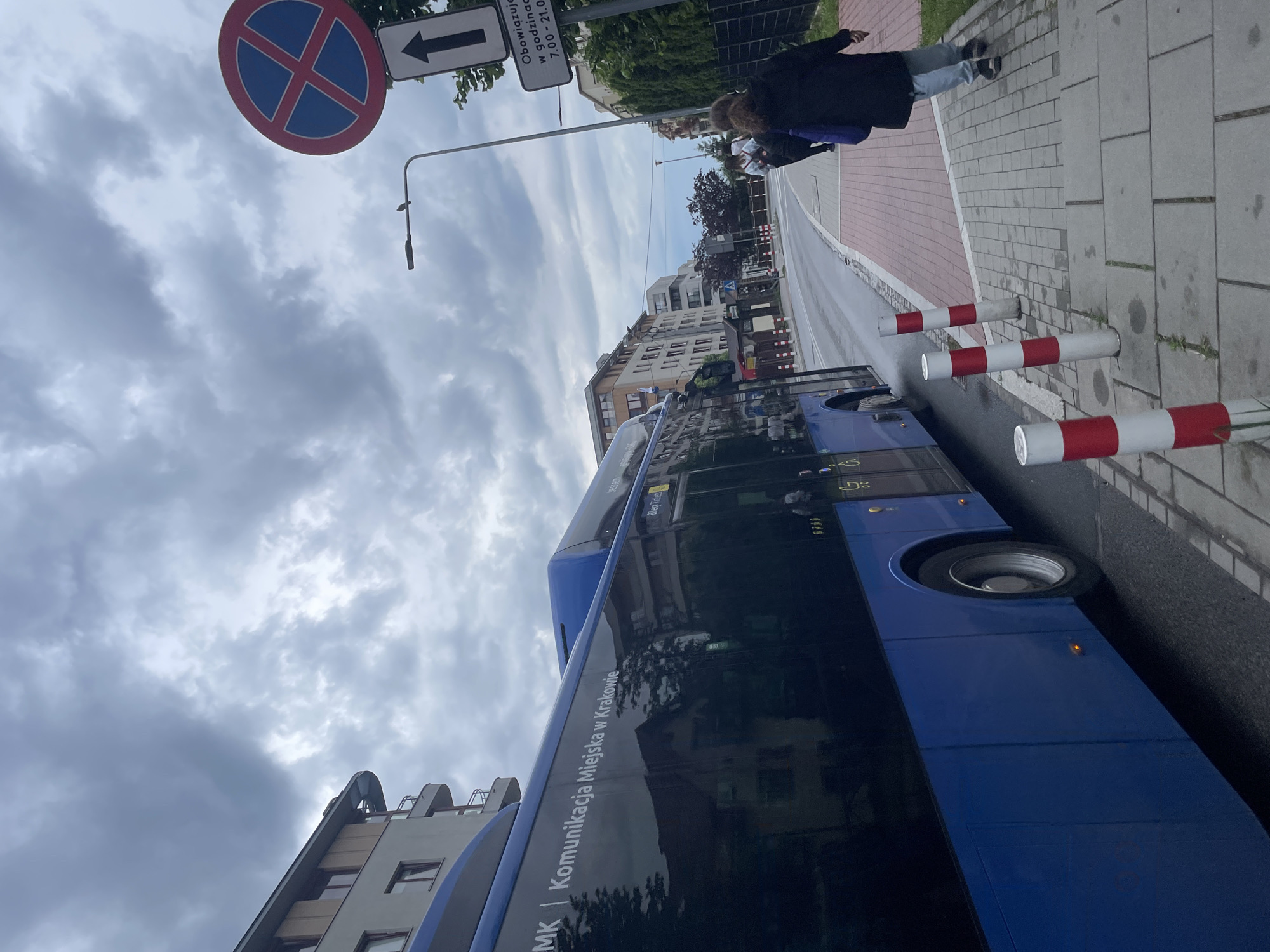 The image, oriented counter-clockwise by 90 degrees, captures an urban street scene under an overcast sky. A blue city bus, part of "KMK | Komunikacja Miejska w Krakowie" (Municipal Transport in Krakow), is prominently visible on the left side, displaying "Piekne Widoki" and partial route numbers like "160" and "06".
To its right, a road leads to a paved sidewalk protected by several red and white striped bollards. A person wearing a dark jacket and light trousers is seen walking away from the camera on the sidewalk. In the background, several multi-story buildings line the street under a cloudy, grey sky.
Overhead, a street light pole supports traffic signs, including a "No Stopping" sign (red circle with a blue cross) and a white sign with a black arrow pointing left. Below the arrow, text in Polish reads: "Obowiązuje w godzinach 7.00 - 21.00" (Applies from 7:00 to 21:00).
The scene depicts everyday urban activity. The bus's livery and the language on the sign identify the location as Krakow, Poland. The time of day appears to be daytime, though the heavy cloud cover prevents strong shadows and suggests a muted light.