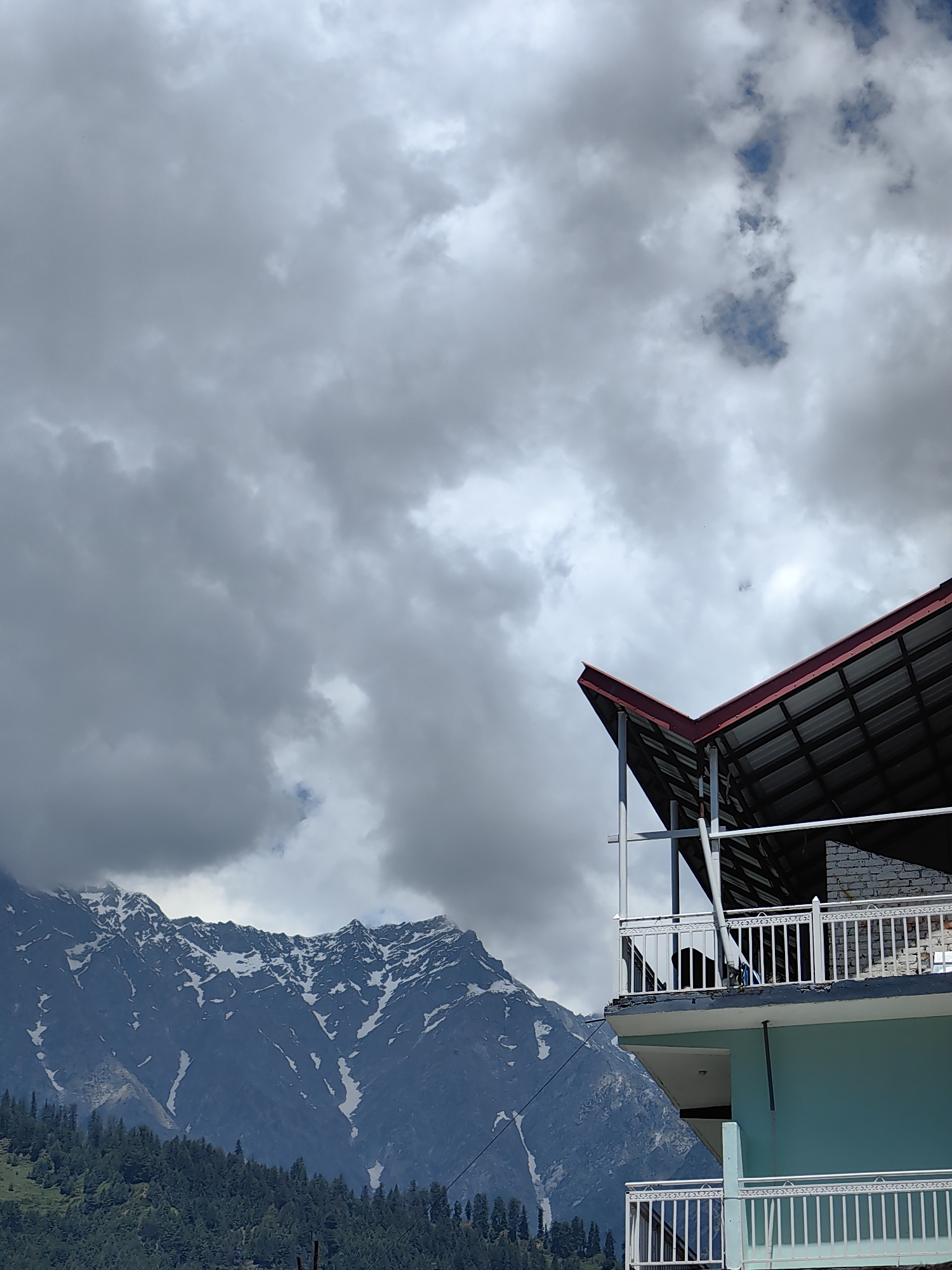 The image captures a partial view of a multi-story building against a backdrop of snow-capped mountains and an overcast sky in Vashisht, India. The sky occupies the upper two-thirds of the frame, dominated by dense, grey cumulus clouds with sporadic patches of lighter blue visible, particularly in the upper right quadrant.
In the mid-ground and background, on the left side, rugged dark blue-grey mountains are prominently featured. Their peaks and upper slopes are marked with significant deposits of white snow. A dense, dark green forest line, composed of coniferous trees, extends along the base of these mountains, transitioning into the lower-left foreground.
On the right side of the image, a segment of a building is visible. Its roof, characterized by a steep angle, is framed in dark red and covered with dark grey or black corrugated sheeting. Below the roof, an upper-level balcony features white vertical metal railings. A dark, shadowed interior space is visible behind these railings, with a brick-textured wall section to the right. The exterior wall of the building's lower visible section is painted a light turquoise or teal color. Another balcony, also with white metal railings, is partially visible at the bottom right of the frame, positioned below the turquoise wall section. No individuals are present in the scene.