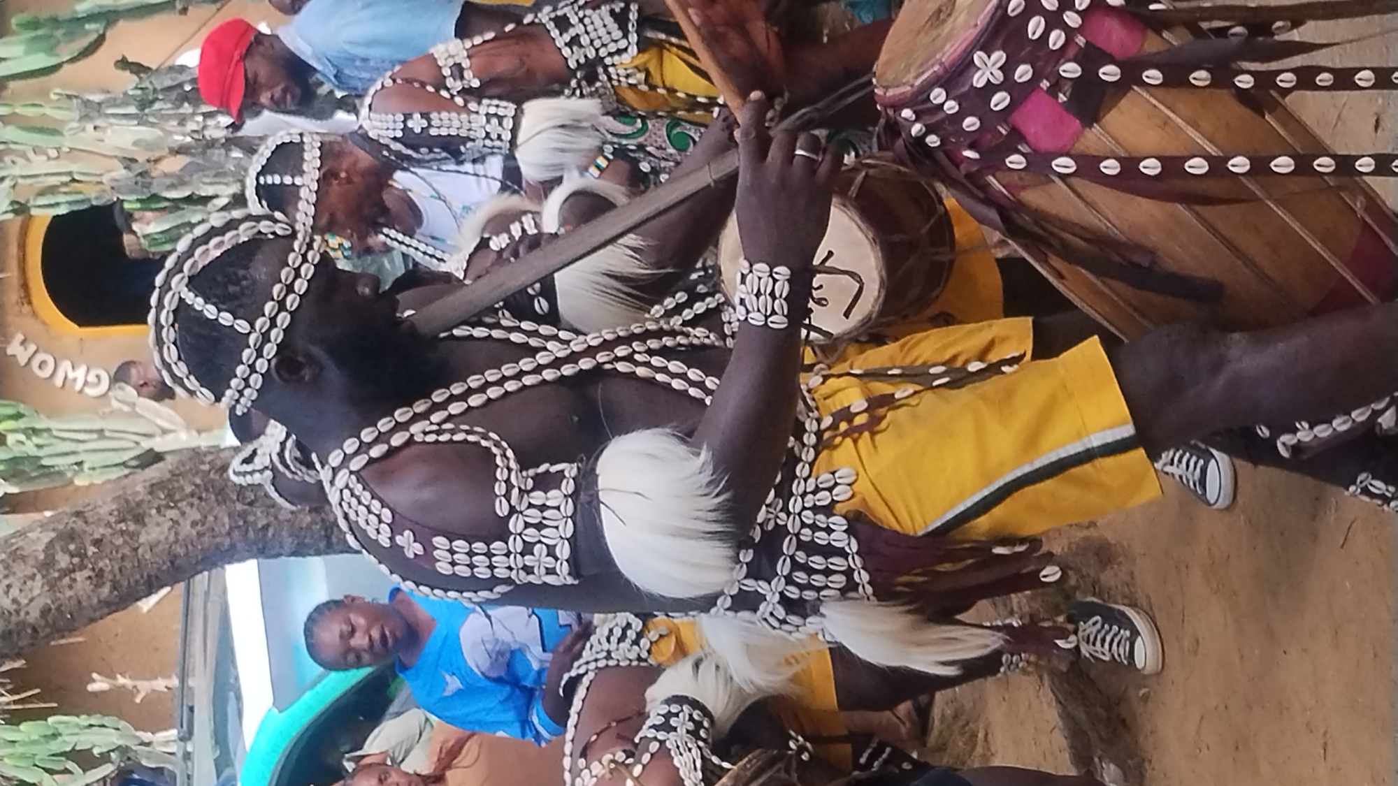 The image depicts a cultural event taking place in Jos, Nigeria. In the foreground, a man adorned in traditional attire featuring cowrie shells and white feather accents is playing a drum. He wears a headdress and a vest decorated with cowrie shells, and yellow shorts. His hands are holding drumsticks, one of which is extended towards a drum positioned in front of him. Behind him, other individuals in similar attire are visible, some also playing drums. In the background, a structure with the word "GWOM" inscribed above an archway suggests the location is a cultural center or a place of significance. A blue car is partially visible on the left side of the frame. The ground appears to be dirt or sand, typical of outdoor settings in Nigeria. The overall atmosphere suggests a lively performance or celebration.