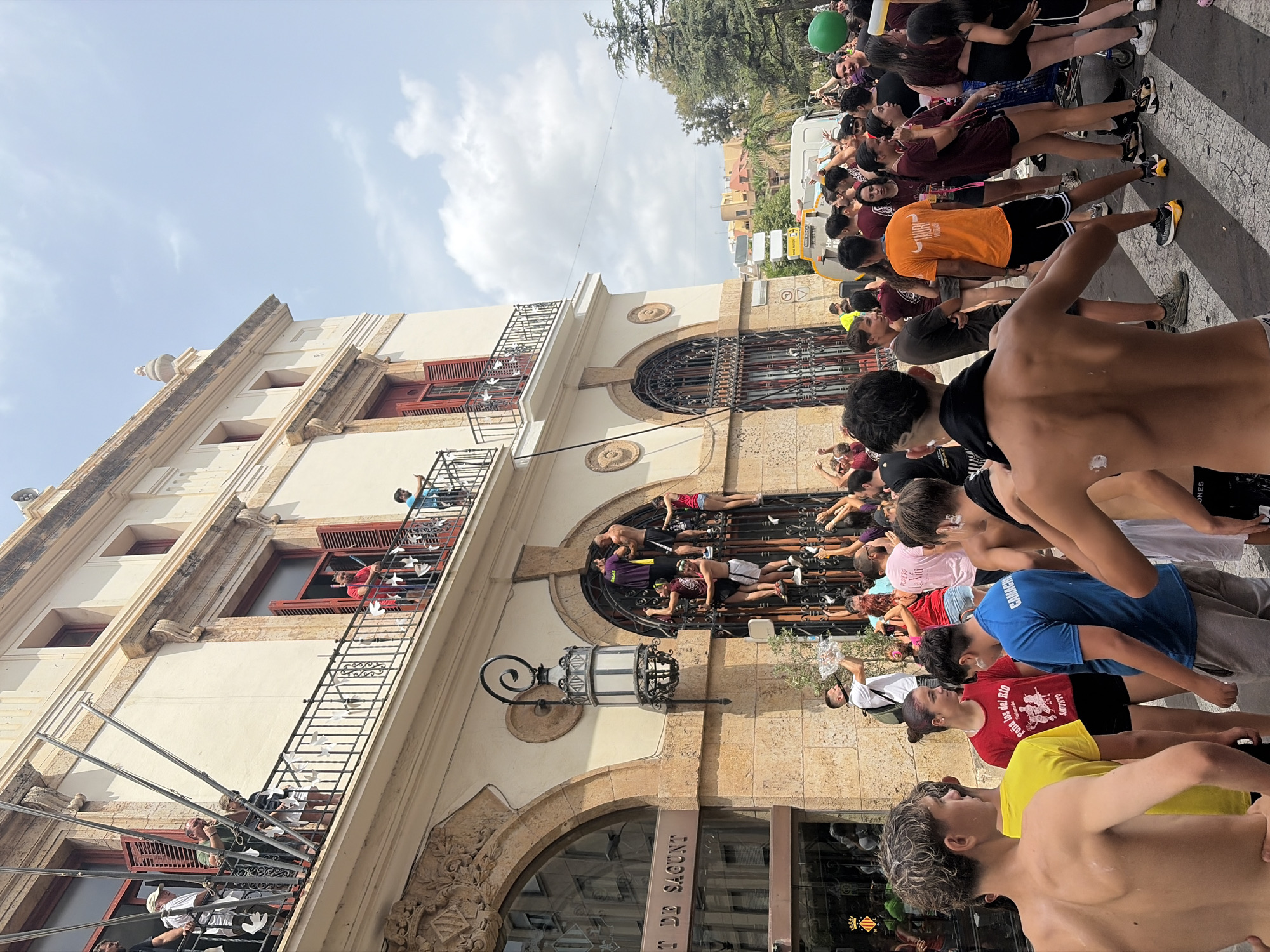 A group of young people are climbing a building in the city of Sagunto, Spain. The building appears to be an old, ornate structure with balconies and large arched windows. The people are dressed in casual clothing, and some are shirtless. They are holding onto each other and the building's facade to climb. The crowd below is watching and cheering them on. The scene appears to be part of a festival or celebration. The sky is blue with scattered clouds.