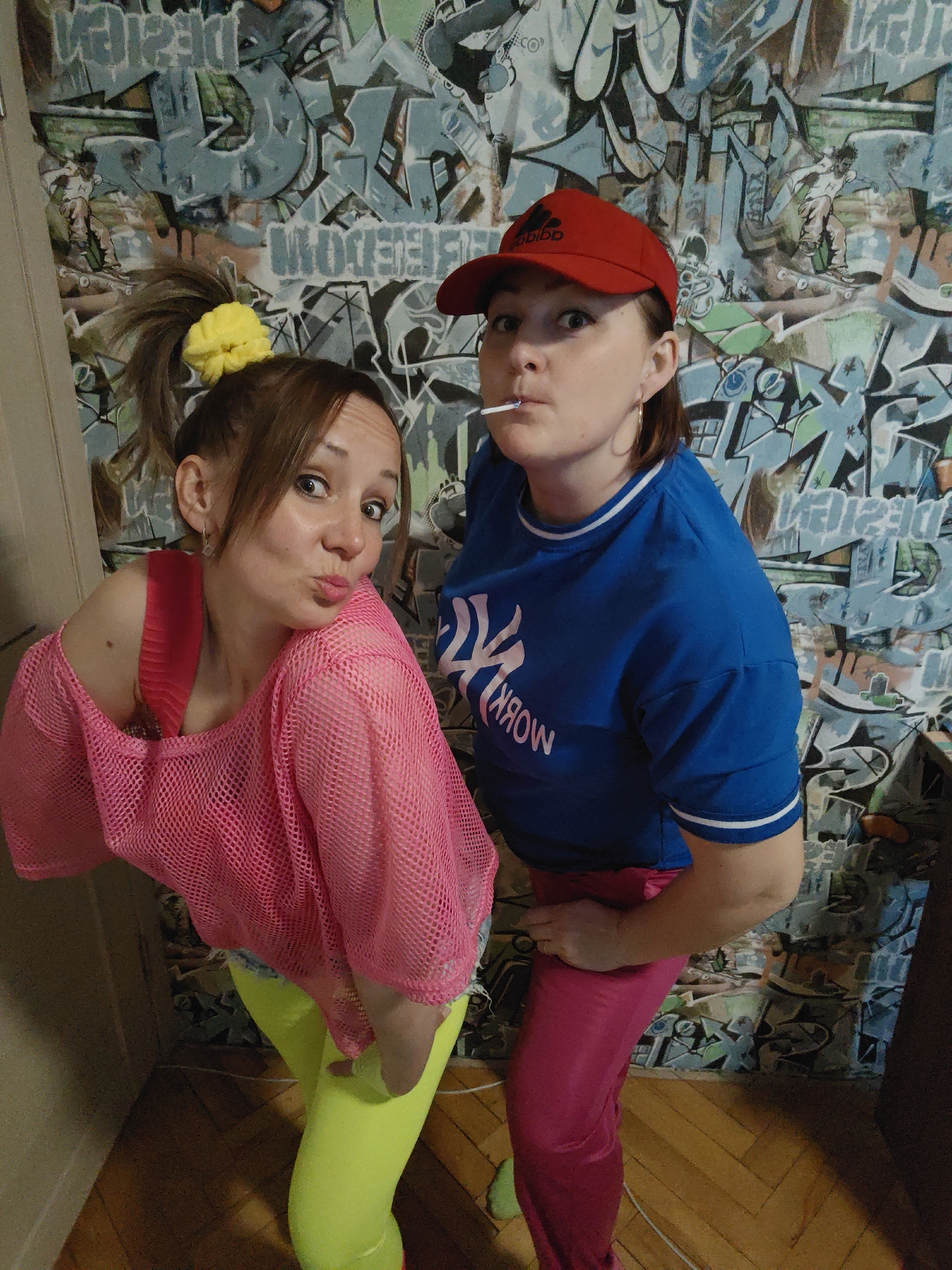 Two women are posing in front of a wall with graffiti. The woman on the left has her hair in a ponytail with a yellow scrunchie, a pink mesh top over a red tank top, and neon green leggings. She is making a kissing face at the camera. The woman on the right is wearing a red Adidas baseball cap, a blue t-shirt with white trim, and pink pants. She is holding a lollipop in her mouth and looking at the camera with a surprised expression. The location is Марковский, Russia.