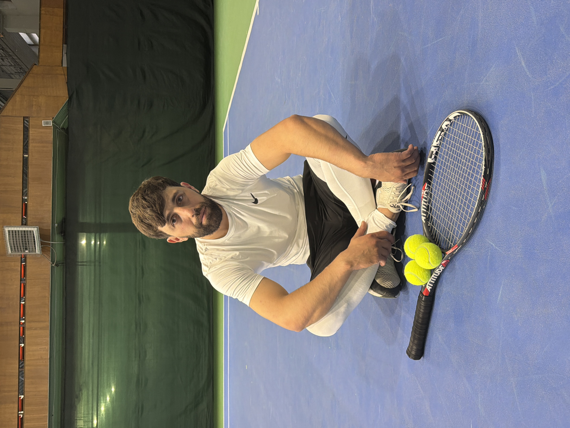 A man with a beard is crouched on a blue and green tennis court. He is wearing a white t-shirt, black shorts, and white leggings. A tennis racket lies next to him, with three tennis balls on its strings. The court appears to be indoors. Based on the 'Attitude' label on the racquet, this may have been taken at the "Tennis Palace Attitude" in Krasnoyarsk, Russia.