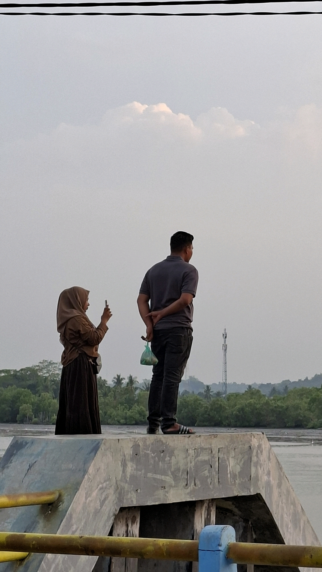 Two individuals stand on a concrete pier overlooking a body of water with a lush, tree-lined shore in the background. A woman, wearing a brown hijab and a long brown dress, is positioned to the left. She is holding a mobile phone in her right hand, appearing to take a photograph. To her right stands a man in a dark grey polo shirt and dark trousers. His hands are clasped behind his back, and he holds a green plastic bag in his left hand. The sky above is overcast with a few wispy clouds. The pier is equipped with yellow railings in the foreground. The scene appears to be the Ujong Blang beach area in Lhokseumawe, Indonesia, a popular spot known for its coastline and recreational activities.