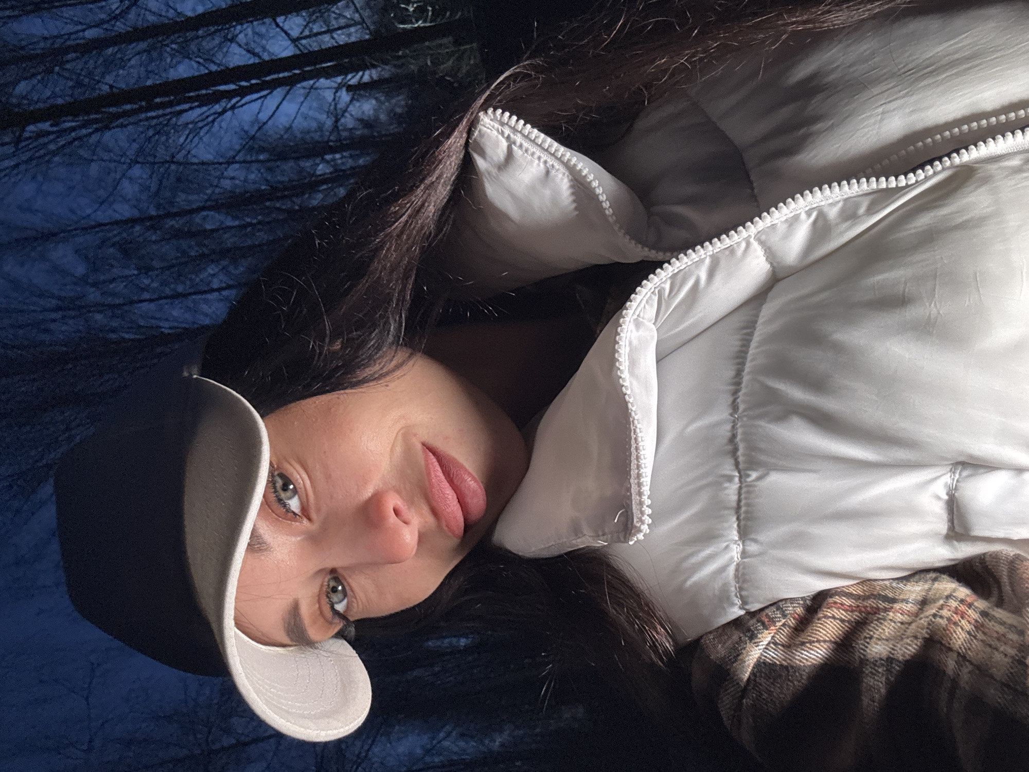 A young woman wearing a white puffer jacket and a dark baseball cap with a light-colored brim lies on her back. She is sticking out her tongue. In the background, there is a dark, blurred view of what appears to be trees against a dusky or nighttime sky. Based on the prompt indicating the location is Chaikovsky, Russia, the backdrop suggests a forest or wooded area near or within the city limits.