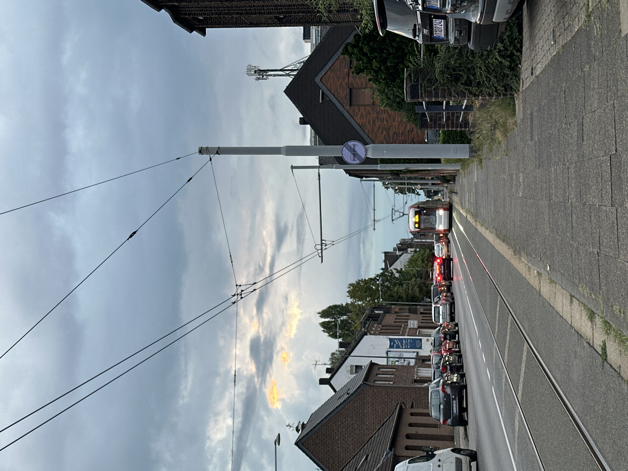 The image depicts a street scene in Krefeld, Germany, on a cloudy day. In the foreground, a paved sidewalk runs parallel to a street with tram tracks. The street is lined with parked cars. Further down the street, a tram is visible, approaching the camera. Buildings, including brick houses and commercial establishments, flank the street. Overhead, power lines for the tram crisscross the sky, and a mobile phone tower is visible in the distance. The sky is overcast with patches of sunlight breaking through the clouds.