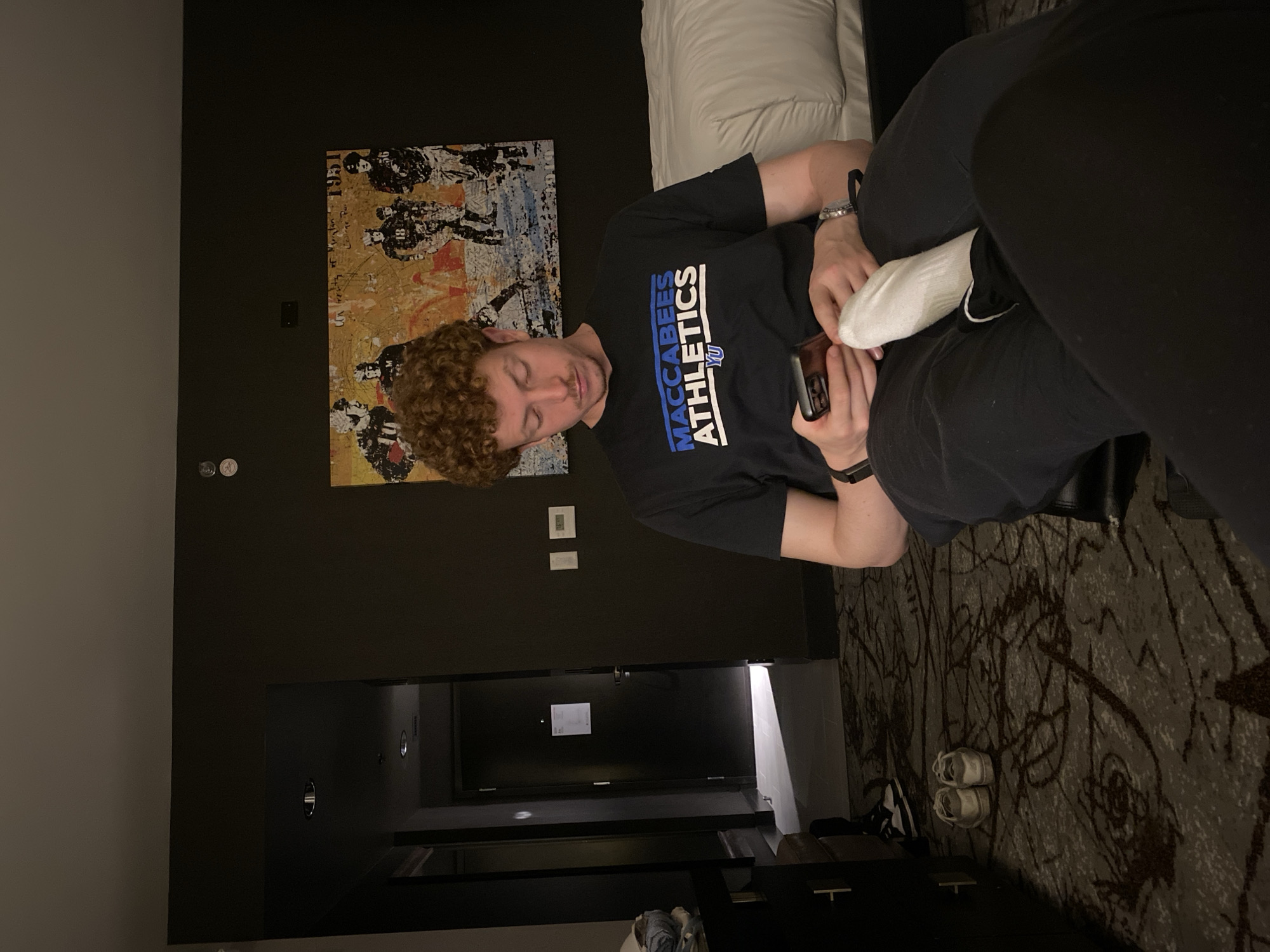 A man is sitting on a bed in a hotel room. He has curly red hair and is wearing a black t-shirt with "Maccabees Athletics" printed on it. He is holding a cell phone in his right hand and a white sock in his left. The man is wearing an Apple Watch on his left wrist and another watch on his right wrist. He is wearing black pants.
Behind the bed is a headboard with white pillows and a black wall with an abstract painting. There is a black door and some electronic items on the wall. The carpet is patterned. There is a dresser and shoes next to the bed.
College Park, United States of America can't be positively identified as the location of this image.