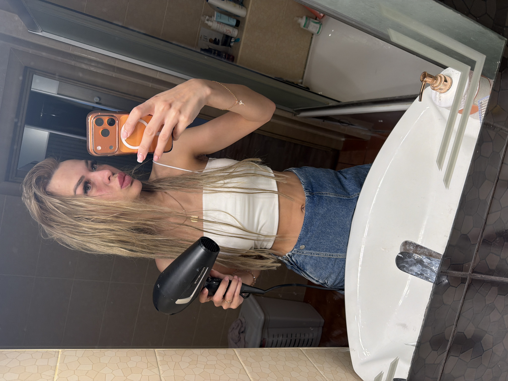 A young woman is taking a selfie in a bathroom. She is holding a black hairdryer in her left hand and an orange-cased smartphone in her right hand, which is angled to capture her reflection in a mirror. The woman has long, blonde, wavy hair and is wearing a white tube top and blue denim shorts. Her makeup appears to be smudged, particularly on her cheeks and forehead. She is posing with her lips pursed.
The bathroom environment is visible in the reflection. To the left, there's a tiled wall with a beige geometric pattern and a grey laundry basket. The sink area in the foreground shows a white sink with chrome fixtures and a soap dispenser with a rose gold pump. The background of the reflection reveals a shower stall with various toiletries on shelves. The overall impression is a domestic setting, likely a personal bathroom. The image does not contain any specific visual cues to pinpoint the location to Kyiv, Ukraine.