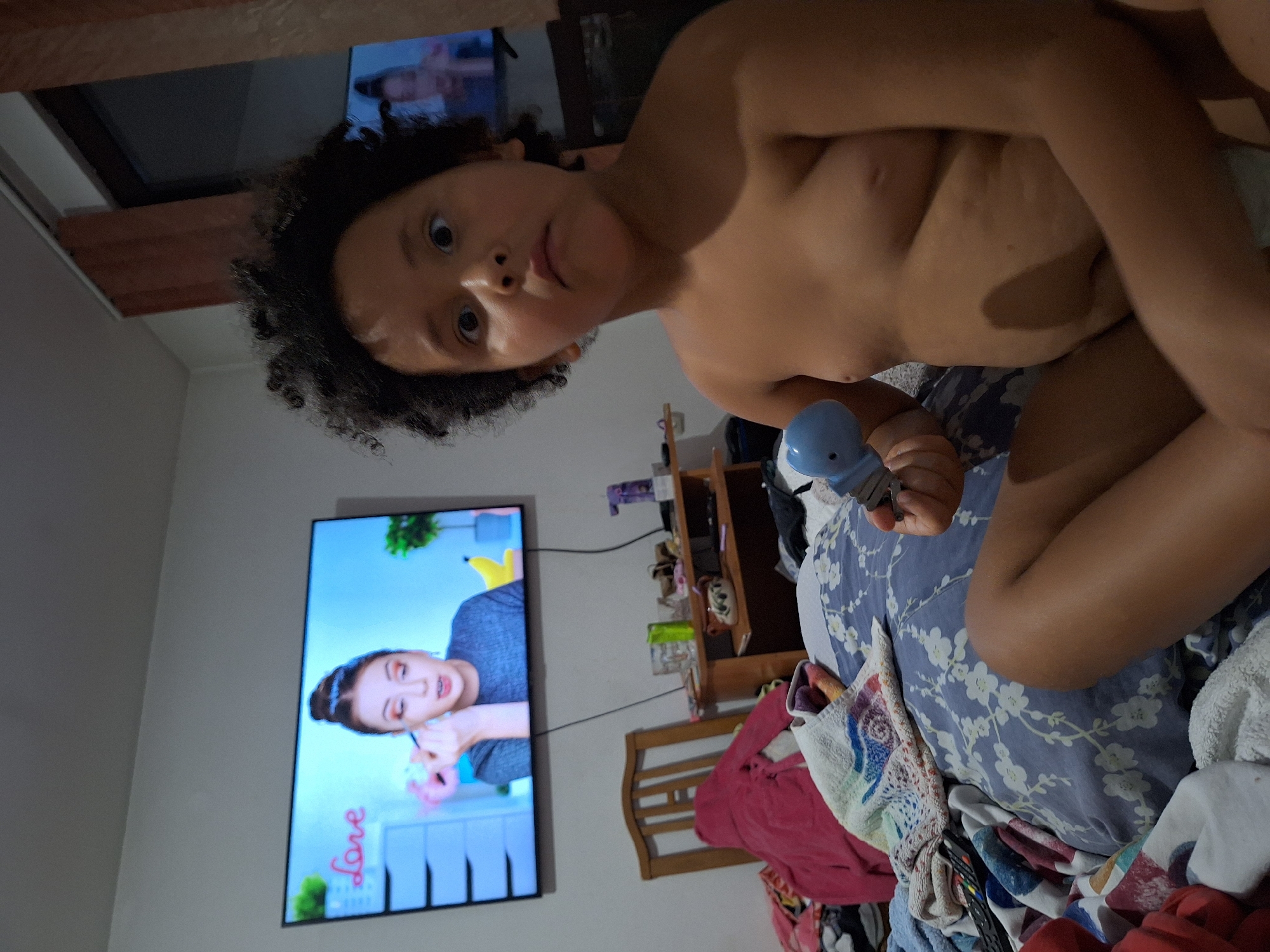 A child with curly dark hair and wide eyes is on a bed, holding a blue toy in their right hand. The child is shirtless. In the background, a television screen displays a makeup tutorial with a woman applying eyeshadow. A wooden chair with a pink robe draped over it is visible to the left of the television. A small wooden table with various items, including glasses and a teapot, sits below the television. The room appears to be a bedroom. The image does not contain any information that would allow for determining its location in Blagoevgrad, Bulgaria.