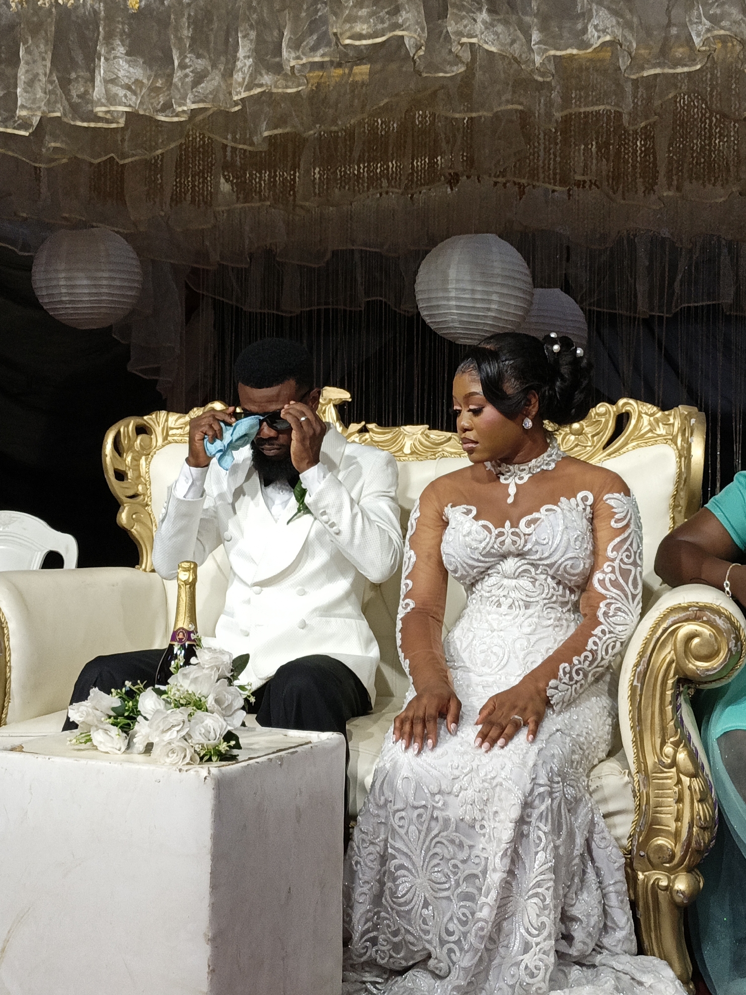 A newly married couple sits on a gold and cream upholstered settee. The groom, on the left, wears a white double-breasted suit over a white shirt and black pants. He has sunglasses on, and he wipes his eyes with a blue handkerchief. To his right, a white cube side table holds a bouquet of white flowers and a bottle of champagne. The bride wears a long, white lace dress and a pearl choker. She has her hair up. The background consists of black curtains and white, spherical paper lanterns. A multi-tiered sheer white drapery hangs above the settee. This photograph was taken in Fagge D2, Nigeria.
