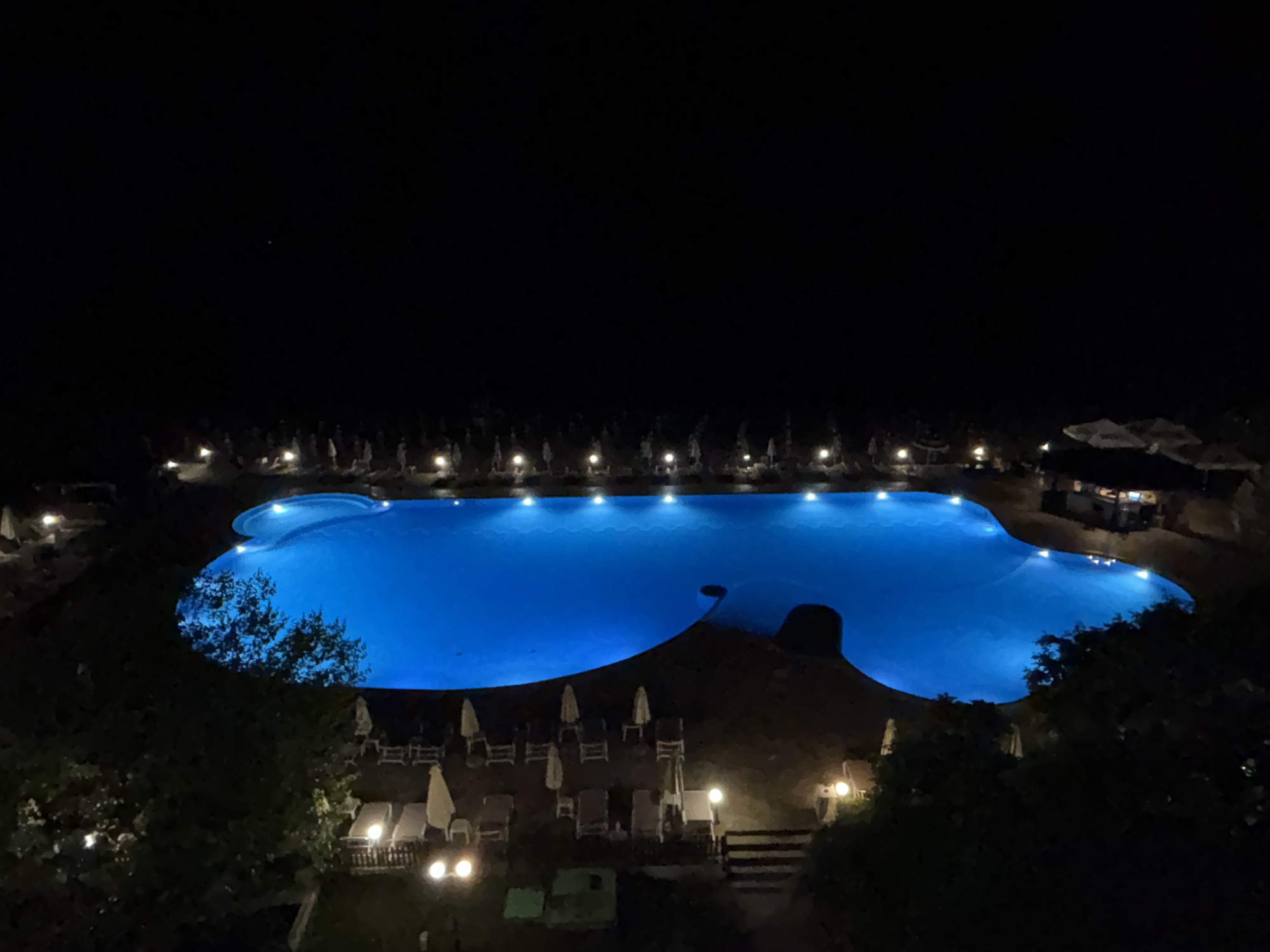 An aerial, night-time, wide shot captures a brightly lit, irregularly shaped swimming pool. The pool's water is a vibrant, electric blue, illuminated by underwater lights. Surrounding the pool, on a sandy or stone surface, are numerous lounge chairs and umbrellas, some of which are also illuminated by nearby lights. The area immediately around the pool is bordered by trees and foliage. In the background, a dark, silhouetted structure, possibly a building or a thatched-roof hut, is visible near the edge of the pool. The sky above is entirely black and devoid of stars or moon. The scene depicts a resort or hotel pool area at night, suggesting it is in a warm climate. The presence of lounge chairs and umbrellas indicates a recreational or relaxation setting. The overall impression is one of a tranquil, inviting nighttime atmosphere. The location is identified as Varna, Bulgaria.