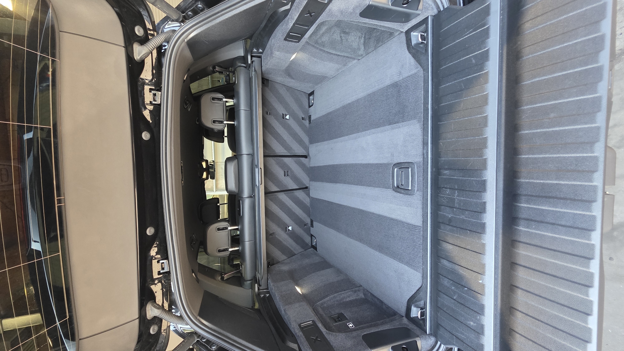 The image shows the open trunk of a black SUV. The interior of the trunk is lined with grey carpeting that has a striped pattern. The rear seats of the SUV are visible and folded down, with black headrests showing. The trunk lid is open and positioned above the interior. The scene is clean and appears to be well-maintained.