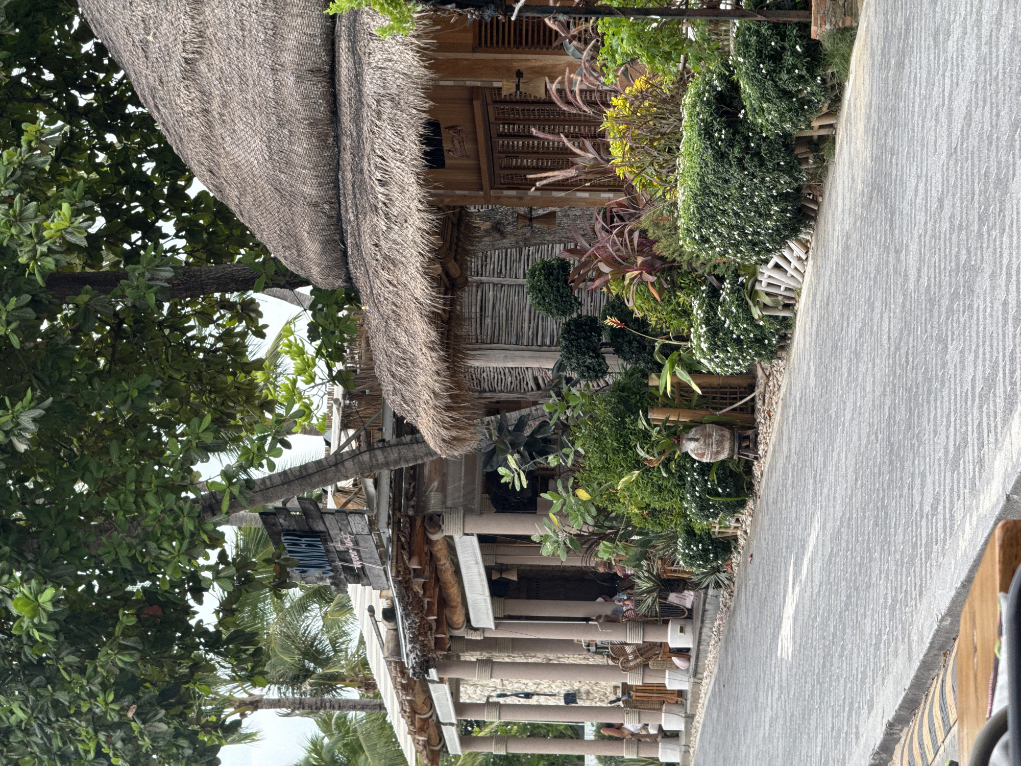 The image depicts a street scene in Ko Samui District, Thailand, featuring a row of tropical-style establishments. On the left, a building with a covered outdoor seating area supported by thick, beige columns is visible. The columns are wrapped with ropes. Above this structure, a weathered wooden sign with the word "SUMMER" in distressed letters is attached to a tree trunk. To the right of this building, a thatched-roof structure with wooden walls and slatted shutters is prominent. Both buildings are adorned with abundant greenery, including lush trees, shrubs, and potted plants. The foreground shows a paved walkway or road with a grey brick pattern. Several people are partially visible inside the building on the left, seated at tables. The overall atmosphere is casual and tropical, suggesting a commercial area with restaurants or shops catering to tourists.
