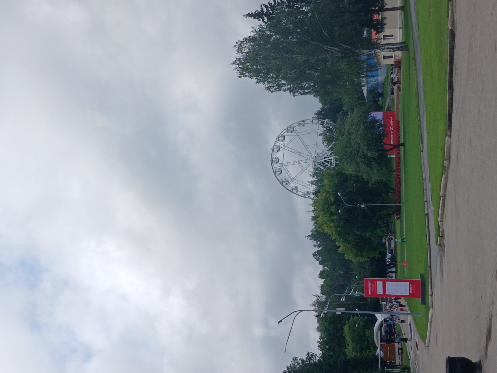 The image depicts an outdoor scene in Yekaterinburg, Russia, likely in an amusement park or a recreational area. In the background, a large white Ferris wheel is prominently visible against a cloudy sky. To the left, a red information kiosk with Cyrillic lettering stands near a paved pathway where several people are walking. Trees and greenery surround the area, and a small blue and yellow building is situated in the middle ground. The foreground shows a paved area with a dark trash receptacle on the left. The overall impression is a slightly overcast day in a public leisure space.