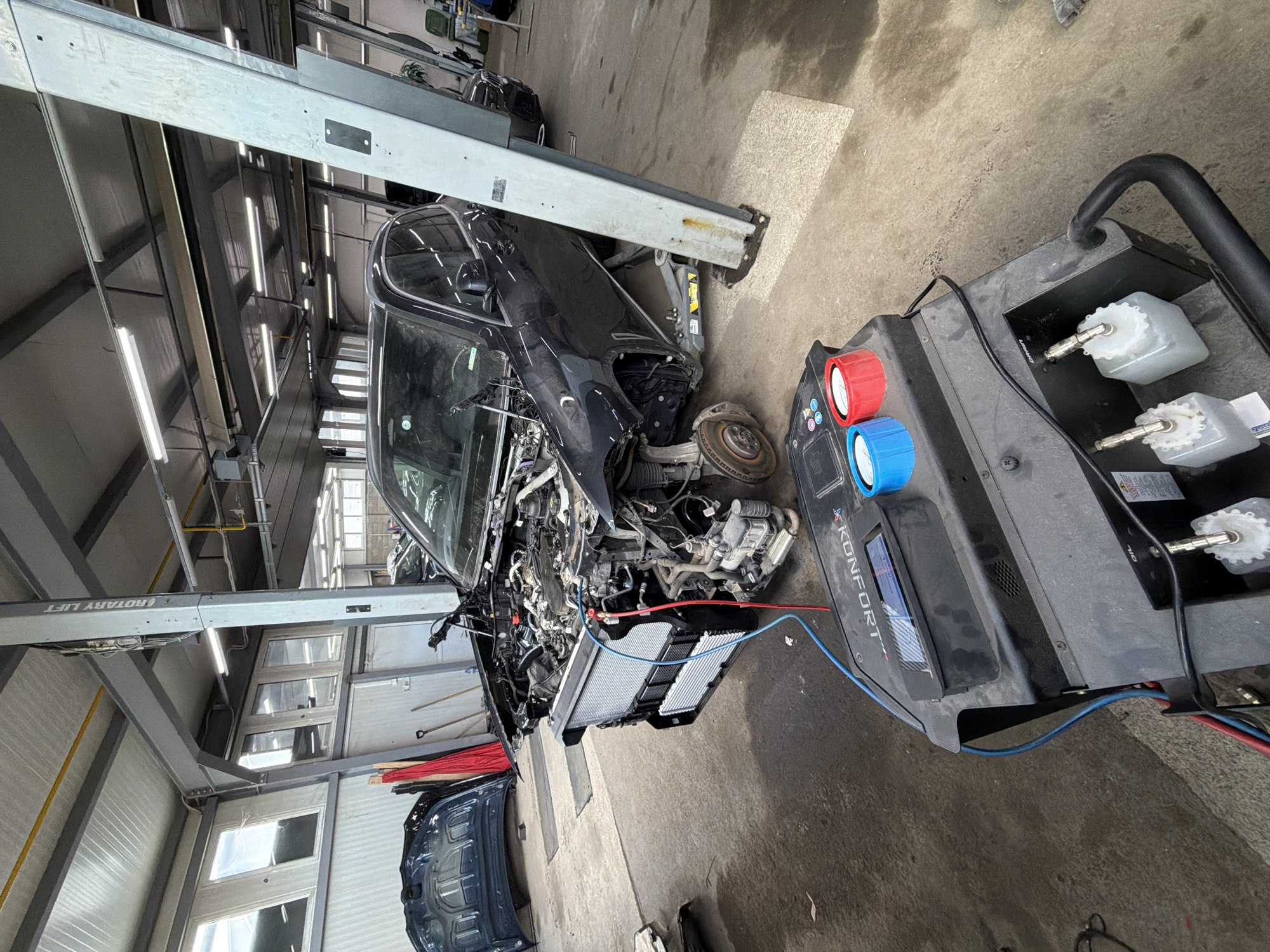 The image shows a car repair shop. A black car is partially disassembled, with its front end exposed and lifted on a hydraulic lift. An A/C recovery machine is connected to the car with hoses. The machine has red and blue knobs and several containers for fluids. The location is identified by the Konfort AC machine which are often sold in Burgas, Bulgaria. The shop has concrete floors, metal supports, and fluorescent lighting. Additional car parts, like a hood, are placed on the floor near the car. The overall impression is that the car is undergoing repairs in a professional automotive workshop.