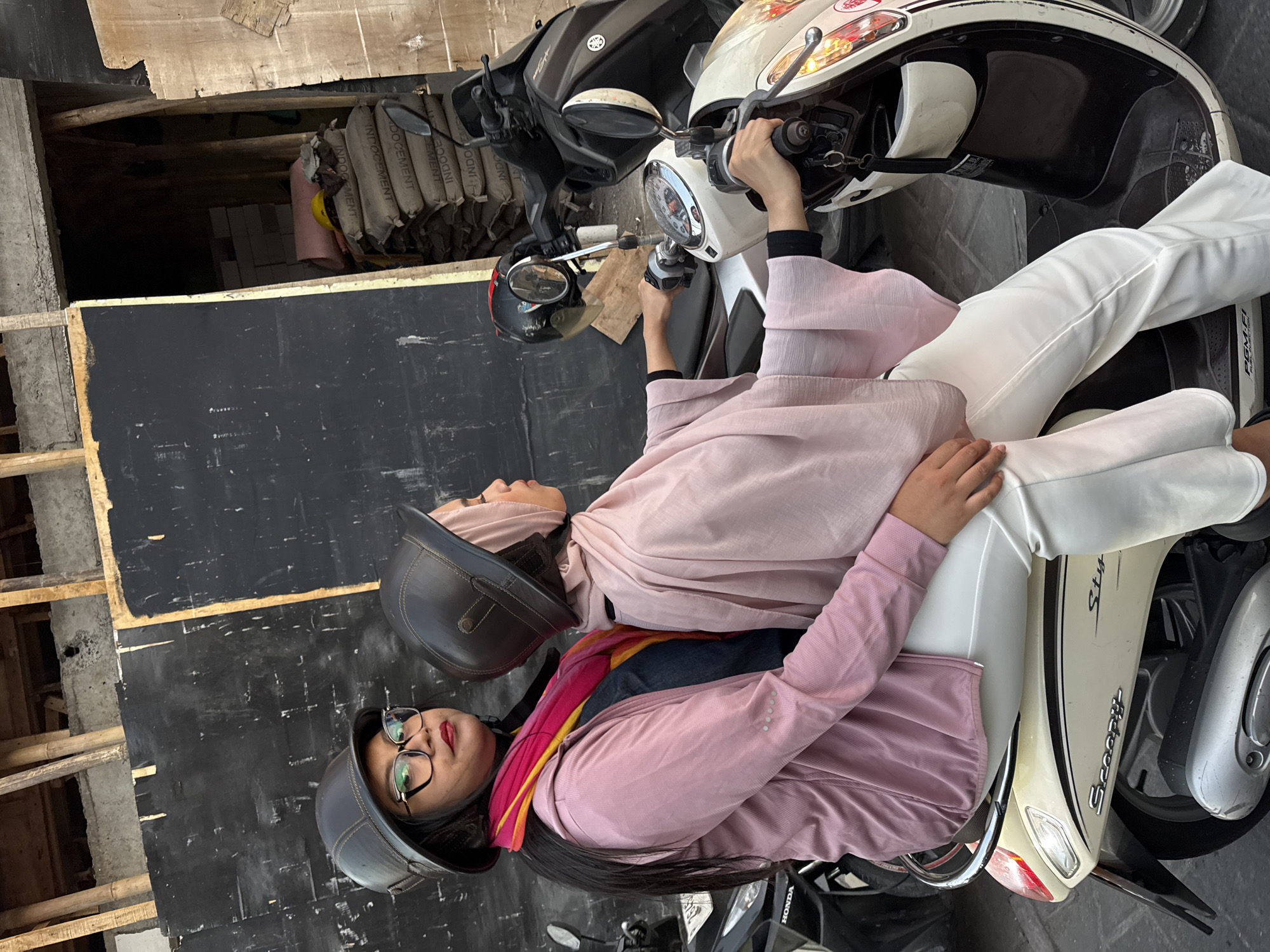 Two women are sitting on a cream-colored scooter, identified by the "Scoopy" logo on its side. The woman in the front is wearing a pink hijab and a pink shawl. The woman in the back is wearing a dark helmet, glasses, a pink jacket, and a colorful scarf around her neck. They are both facing towards the right of the image. The scooter is parked in front of a black wall, with stacks of cement bags visible behind it. The environment appears to be an urban setting in Kuta, Indonesia, possibly a construction area given the presence of the cement bags and unfinished building structures.