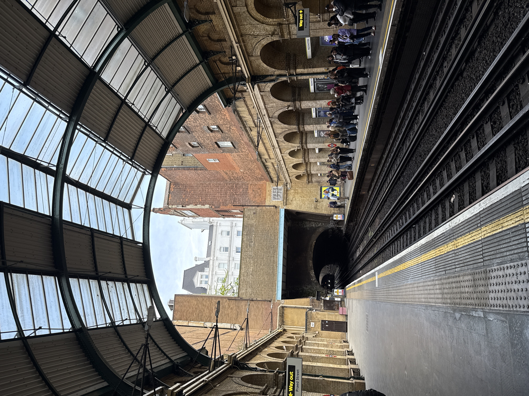 The image shows Paddington Station in London, UK, as seen from an elevated perspective above the platform. On the right side of the frame, train tracks stretch into a tunnel, paralleled by a platform where a crowd of people stand waiting. Above them, arched brickwork forms part of the station's architecture. To the left, a curved glass and steel canopy covers another section of the platform, framing a view of nearby buildings, some brick, others painted white. The overall composition is oriented vertically. Signage indicating "Way Out" and "Praed Street" is visible, confirming the location.