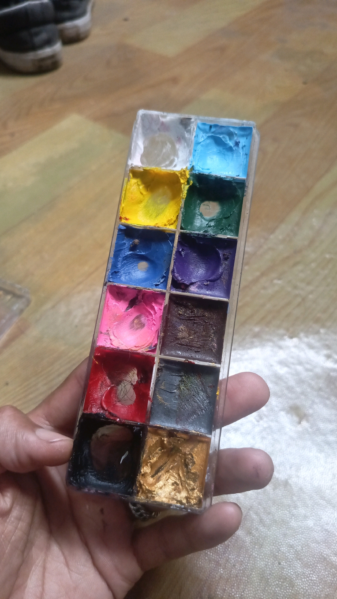 A hand holds a rectangular plastic palette containing twelve compartments filled with colorful paints. The palette is oriented at an angle, with the paints arranged in three rows and four columns. From top to bottom and left to right, the colors appear to be: white, light yellow, light blue, dark green, yellow, blue, purple, pink, dark brown, red, gray, and gold. The paints are textured, suggesting they are oil-based or cream paints, possibly for makeup or art. In the background, out of focus, are the backs of two dark-colored shoes with white soles, resting on a wooden floor. The overall setting appears to be indoors, possibly a domestic environment. There is no specific indication of the city of Kyereku, Nigeria, in the image.