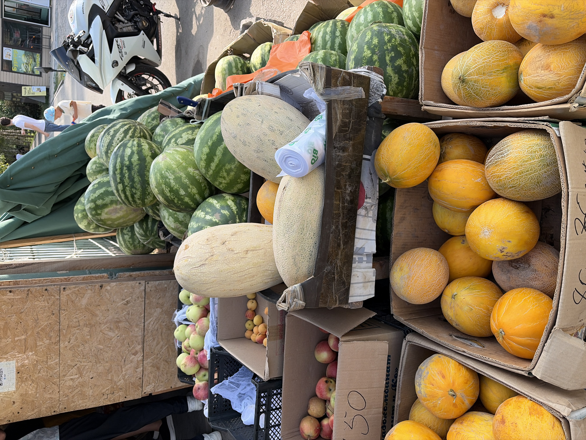 A stall at an outdoor market displays a variety of fruits, including watermelons, cantaloupes, and apples. In the foreground, several cardboard boxes are filled with different types of melons. One box contains mostly yellow-skinned cantaloupes, while another holds a mix of yellow and orange melons. To the left, a box of red and green apples is visible. Behind these, larger melons, including some pale yellow cantaloupes and a large pale green melon, are arranged. In the background, a stack of watermelons is prominently displayed. A white motorcycle is parked to the right of the stall, and a few people are visible in the background, including a child and two adults. The scene appears to be outdoors, with natural lighting.
