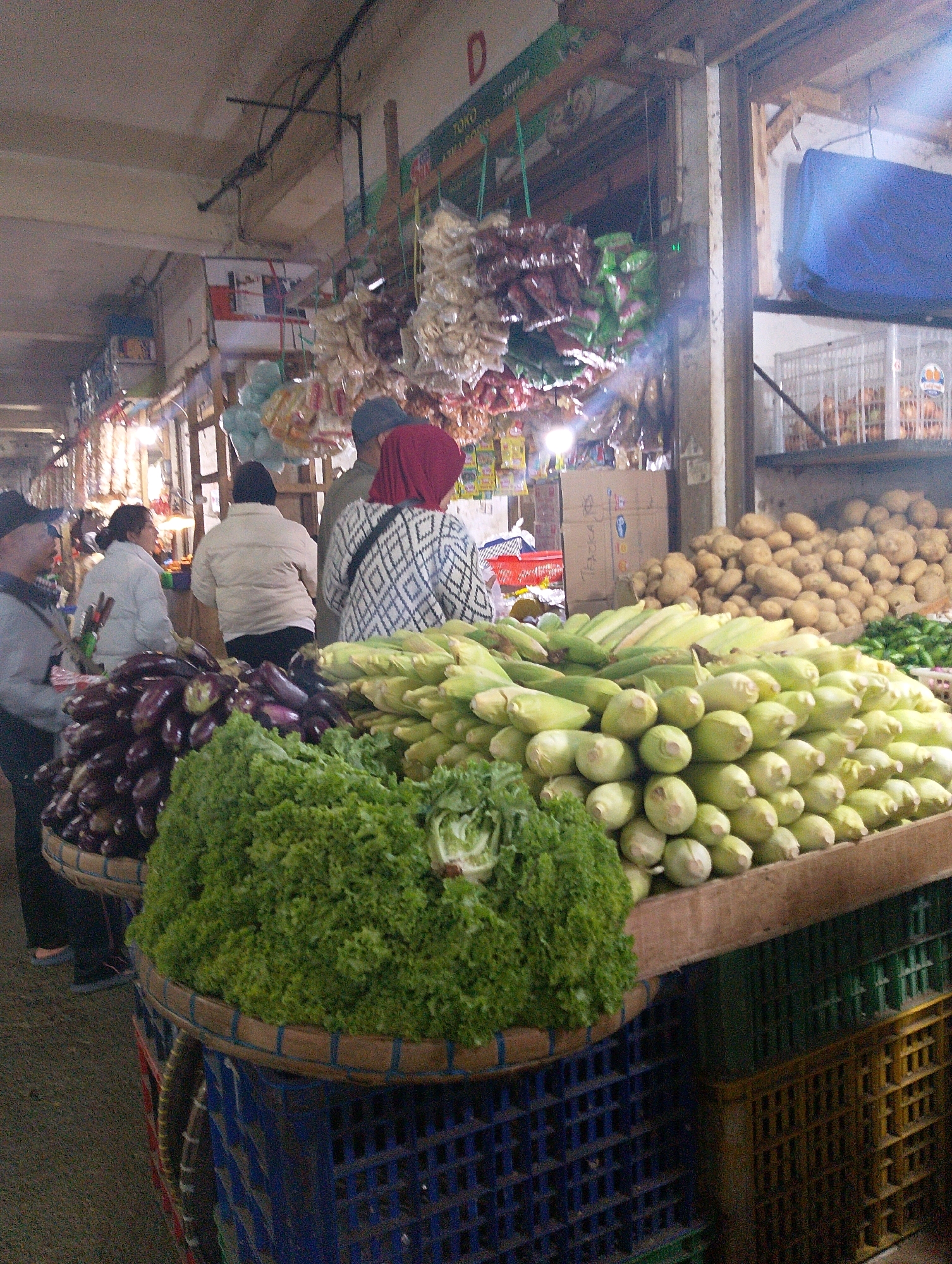 The image depicts a bustling outdoor market scene in Lembang, Indonesia, likely a vegetable and produce market. In the foreground, a large basket overflowed with vibrant green lettuce, and another basket held a generous pile of dark purple eggplants. To the right, a substantial quantity of corn cobs, still in their husks, were arranged on a wooden counter. In the background, a large pile of potatoes was visible, and various dried goods, possibly spices or snacks, were hanging from the ceiling. Several individuals were present, mostly seen from the back or side. One person in the foreground wore a black cap and a grey jacket, facing away from the camera and towards the produce. Another individual in a red headscarf and a patterned top was also visible, seemingly interacting with someone or observing the market. The market environment appears to be covered by a roof, indicated by the ceiling structure and lighting. The overall atmosphere suggests a typical local market with abundant fresh produce on display for sale.